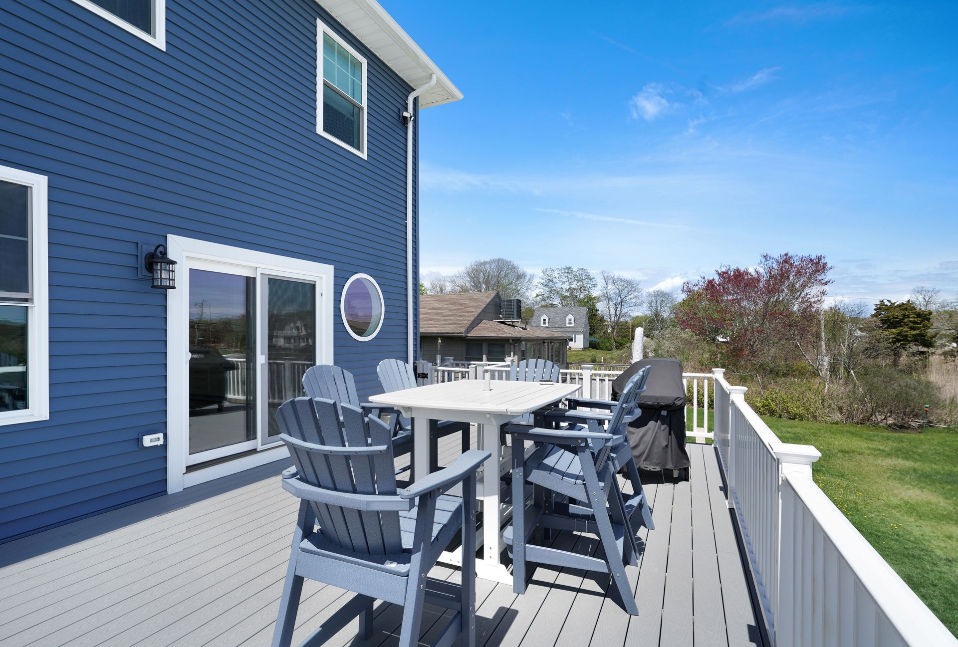 Blue house with a deck featuring a table and chairs, with a view of a yard and sky.