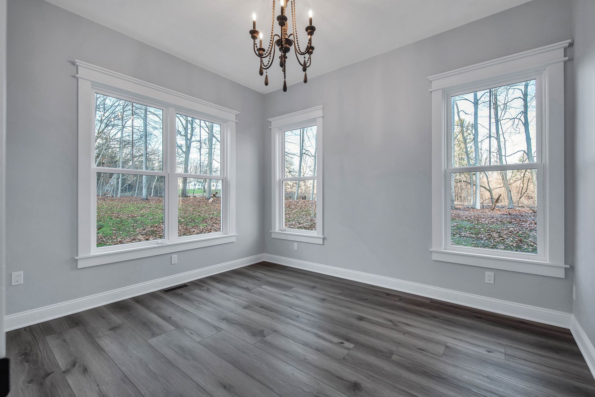 Empty room with three windows, gray walls, dark wood floor, and chandelier.