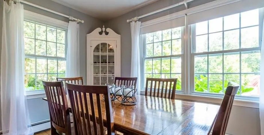 Dining room with wooden table and chairs, corner cabinet, and large windows with sheer white curtains.