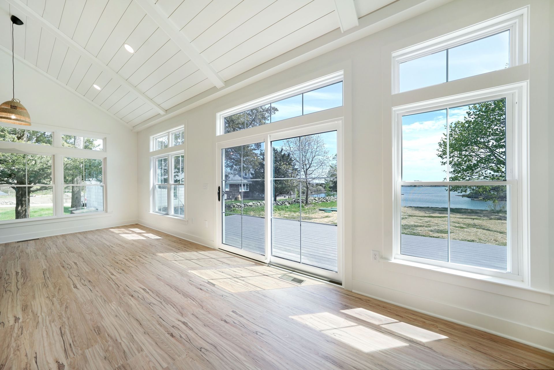 Bright, empty room with large windows overlooking a scenic waterfront. Wooden floor and white walls.