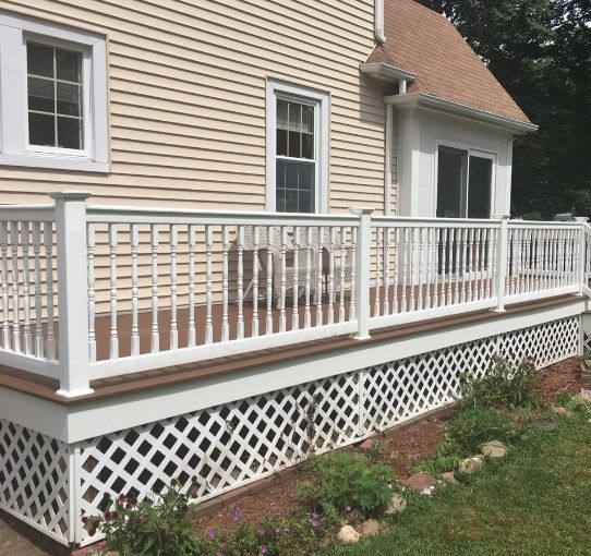 White railing on a wooden deck attached to a light-colored house. Lattice skirting, green grass, and plants surround.