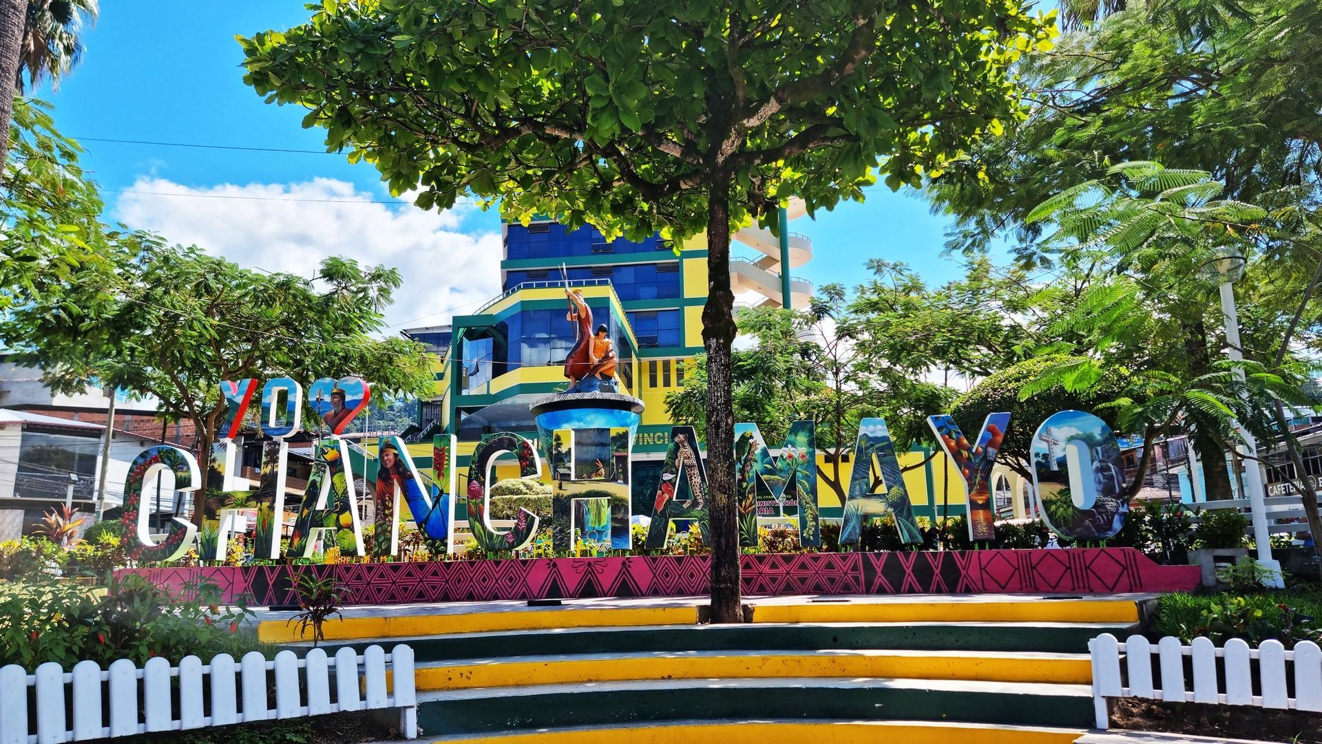 Carretera de montaña que discurre entre exuberantes acantilados verdes bajo un cielo azul despejado, con una señal de tráfico verde en lo alto.