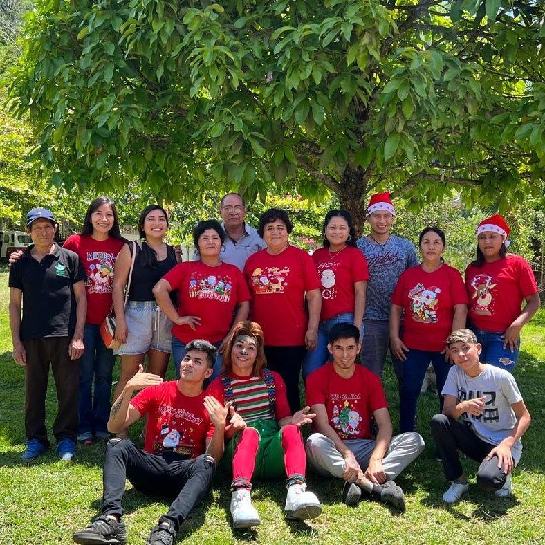 Un numeroso grupo de personas con camisetas rojas con motivos navideños se reúne bajo un frondoso árbol .