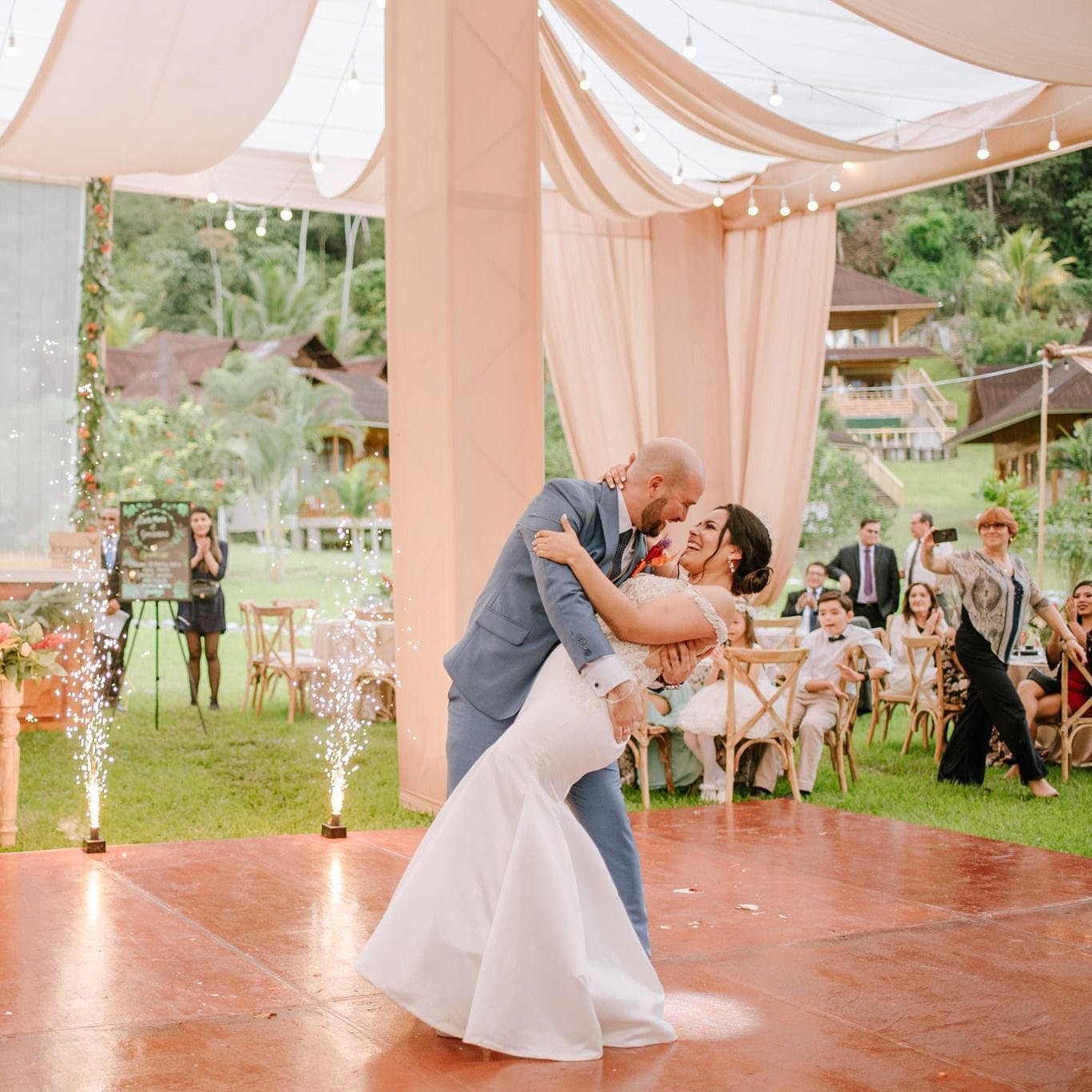 Una pareja bailando durante la recepción de su boda al aire libre, con fuentes resplandecientes en el césped
