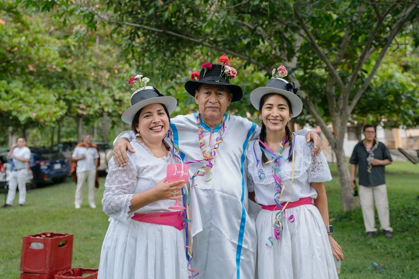 Tres personas vestidas con trajes tradicionales blancos de festival y sombreros adornados con flores sonríen juntas.
