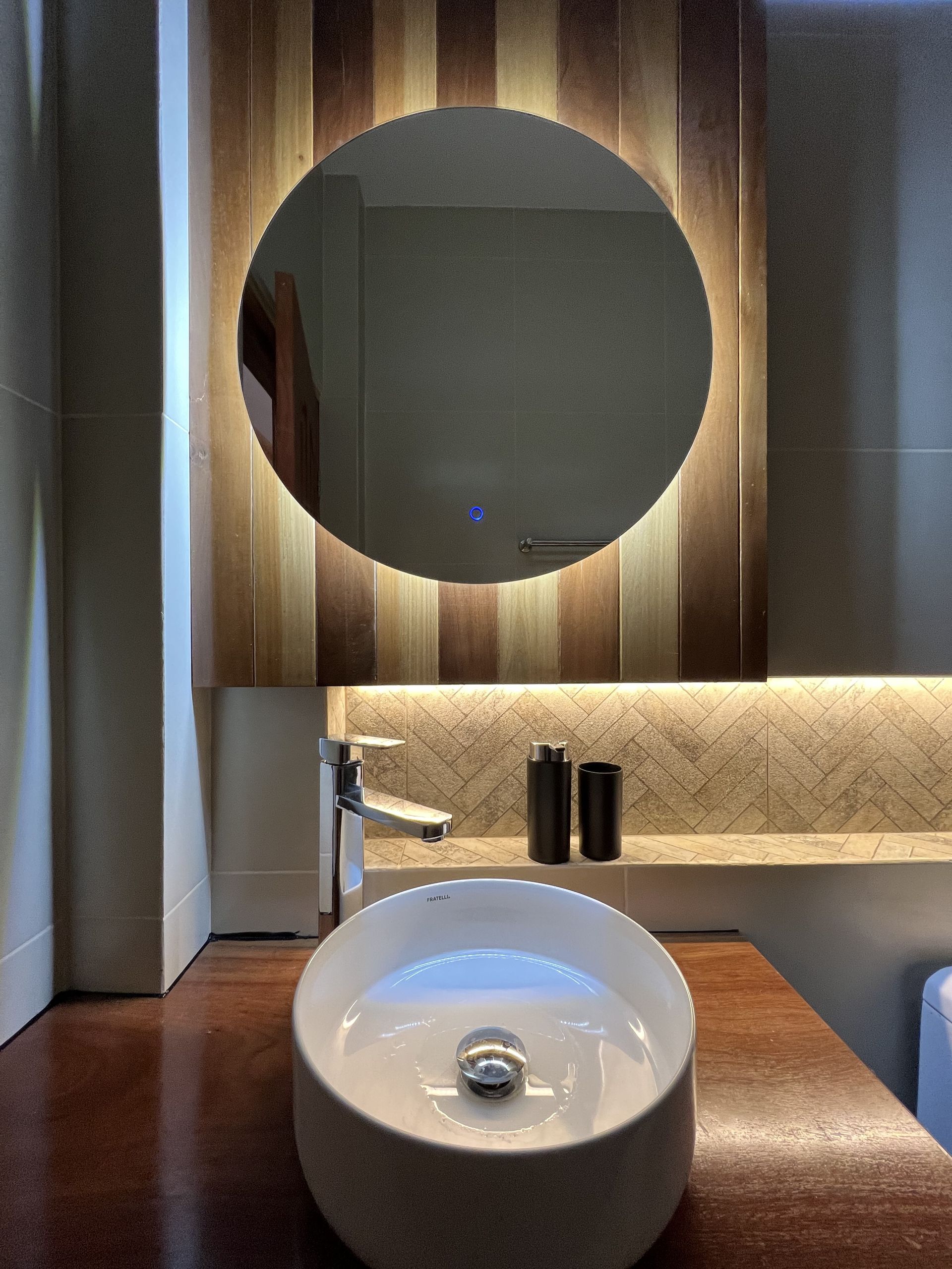 Un lavabo moderno con espejo redondo LED sobre una pared vertical revestida de paneles de madera 