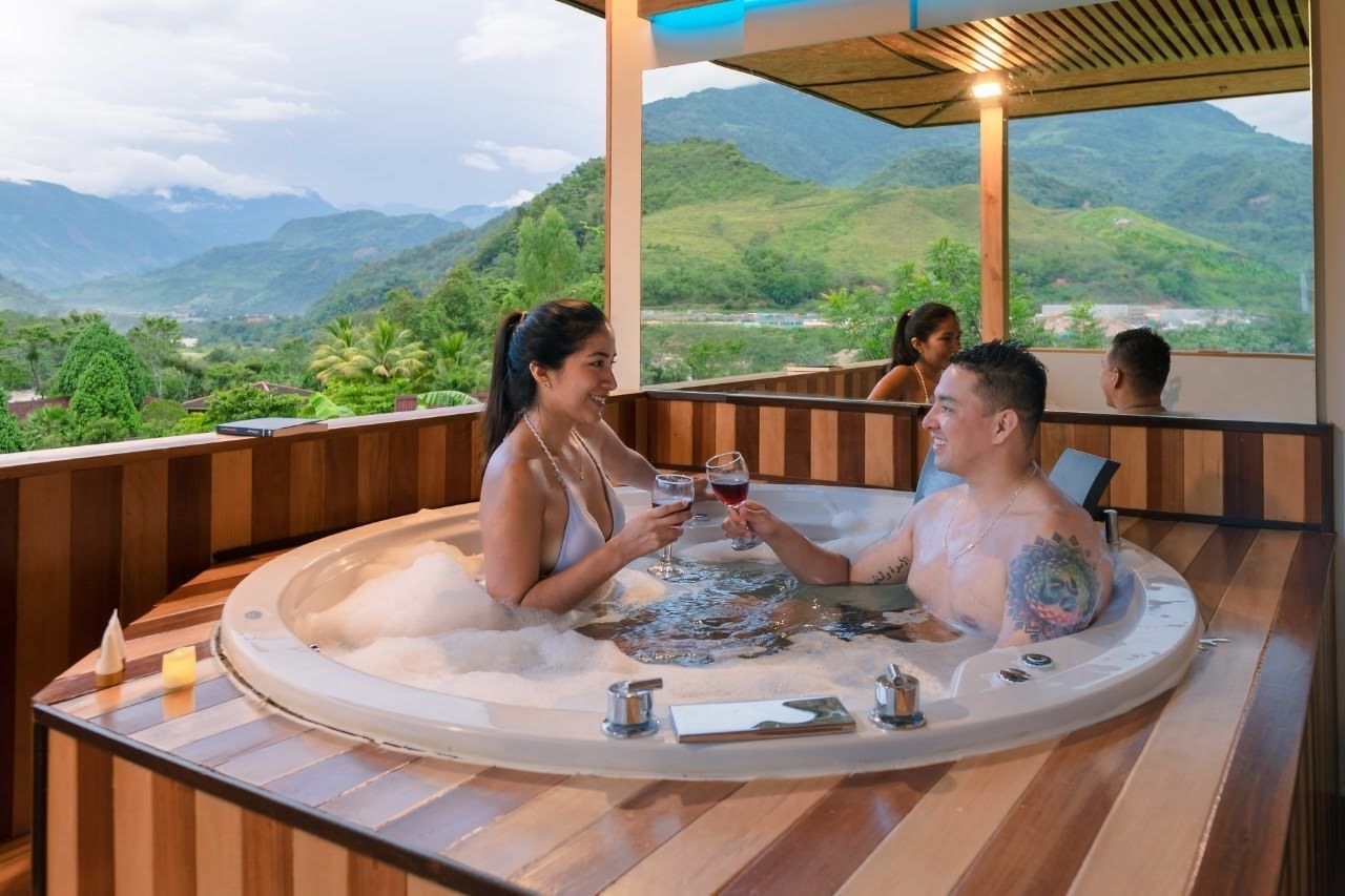 Una pareja brinda con vino en un jacuzzi con vistas a la montaña. Otras personas al fondo.