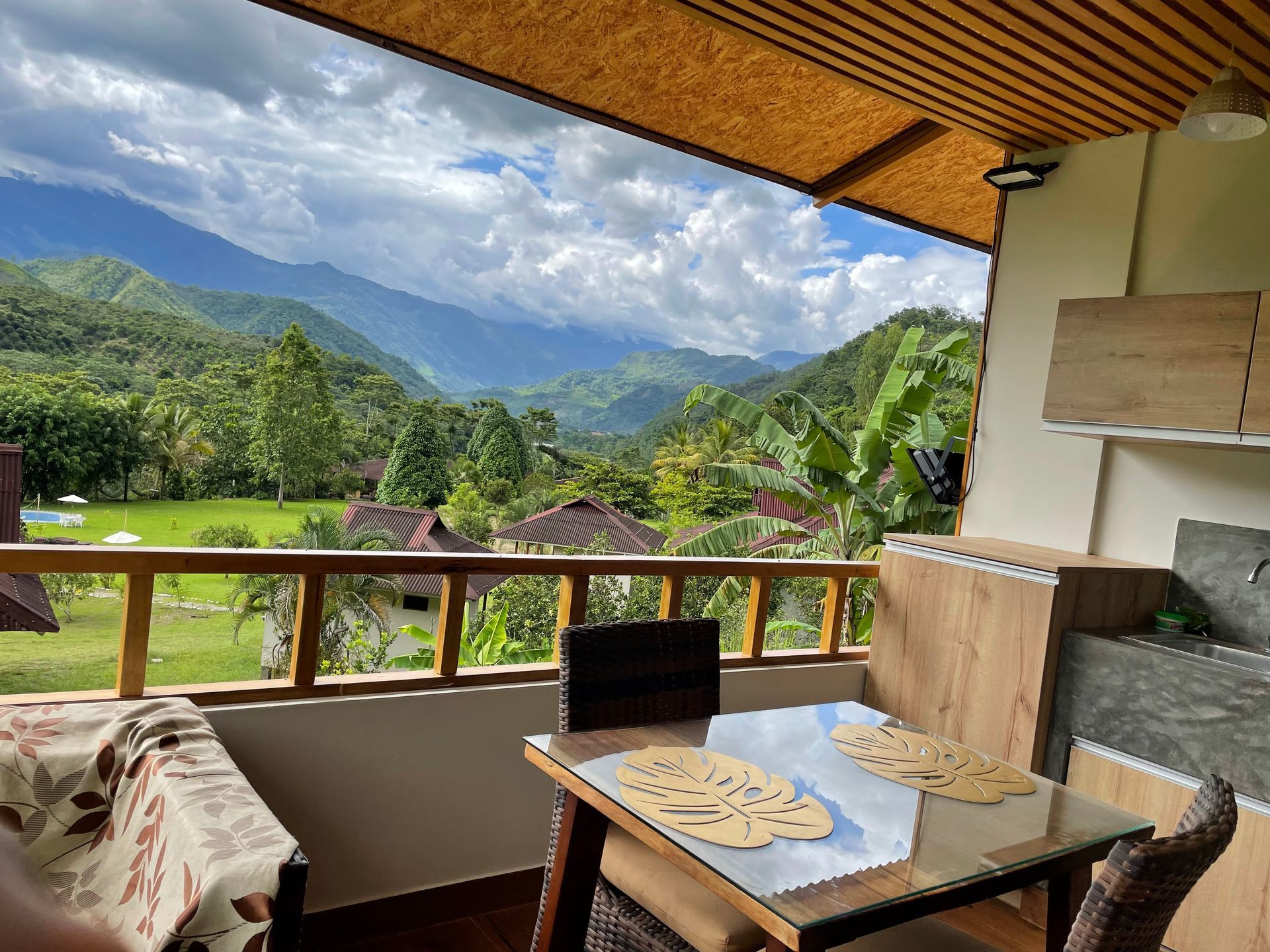 Un balcón con mesa y sillas con vistas a un valle verde exuberante con montañas onduladas bajo un cielo nublado.