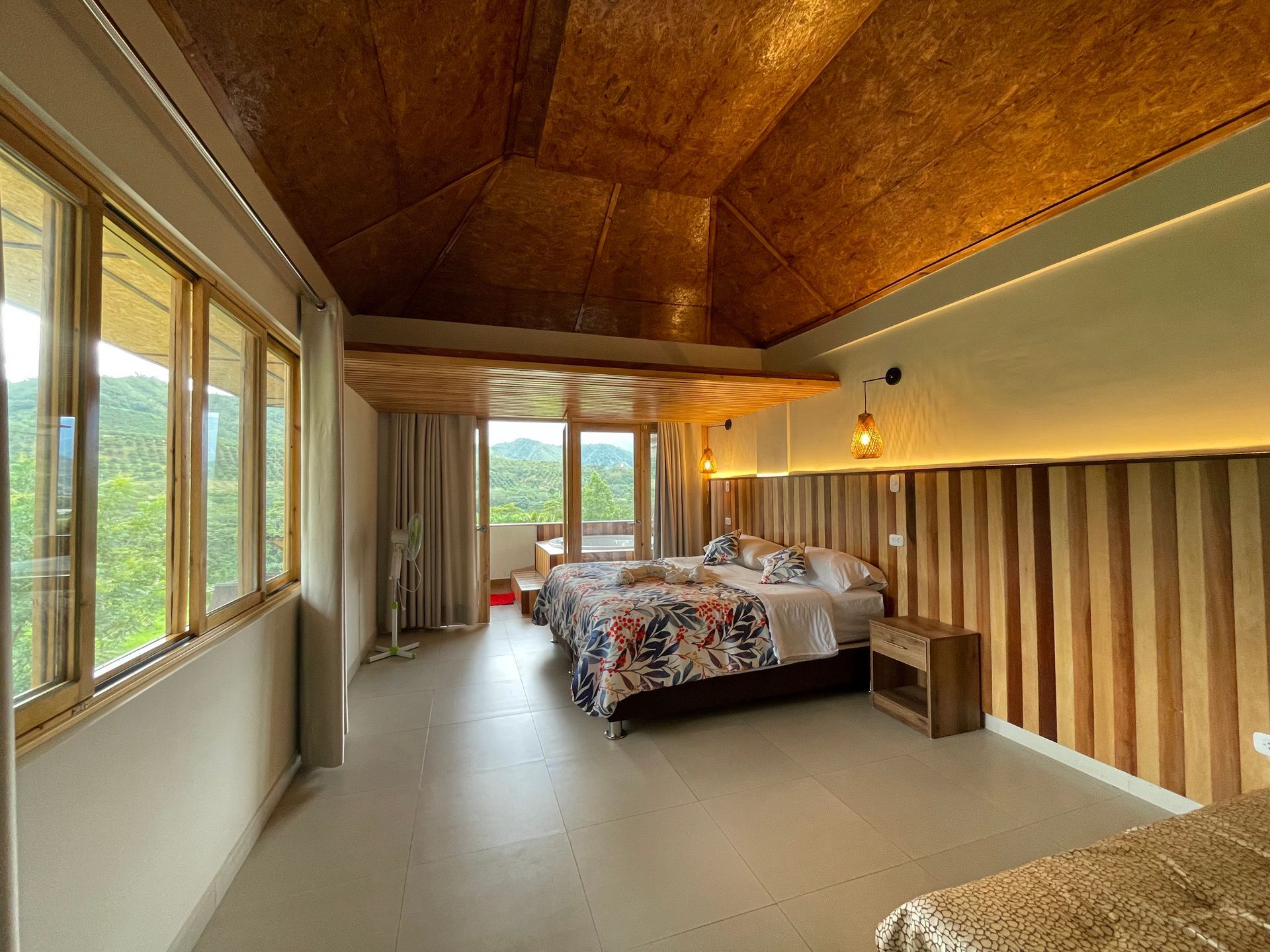 Dormitorio con detalles en madera, grandes ventanales con vistas a un exuberante paisaje y un cubrecama floral.