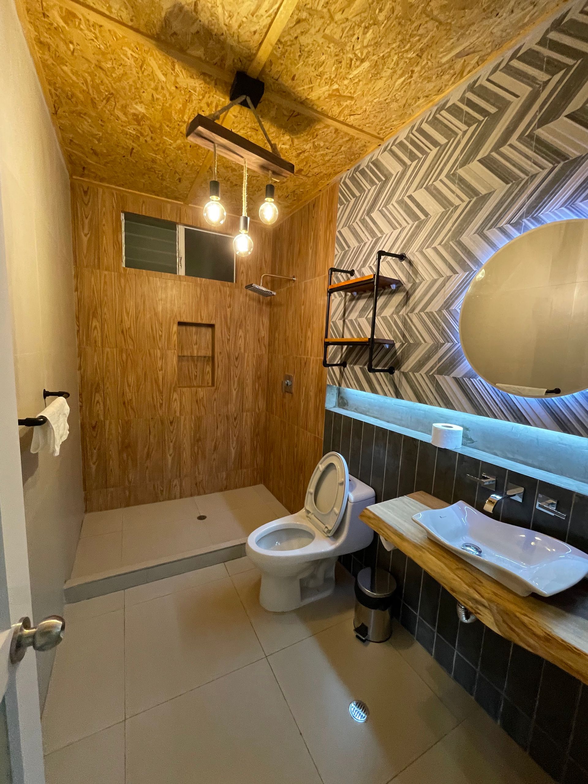 Baño con paneles de madera, pared estampada, inodoro, lavabo y ducha. Lámpara de Edison.