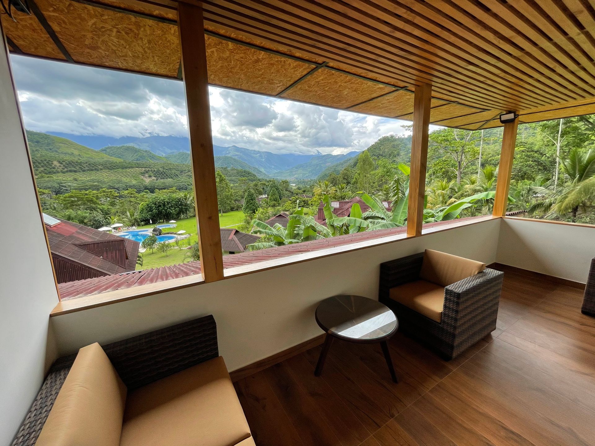 Balcón con vistas al valle, con dos sillas, una mesa y una piscina. Frondas verdes y cielo nublado.