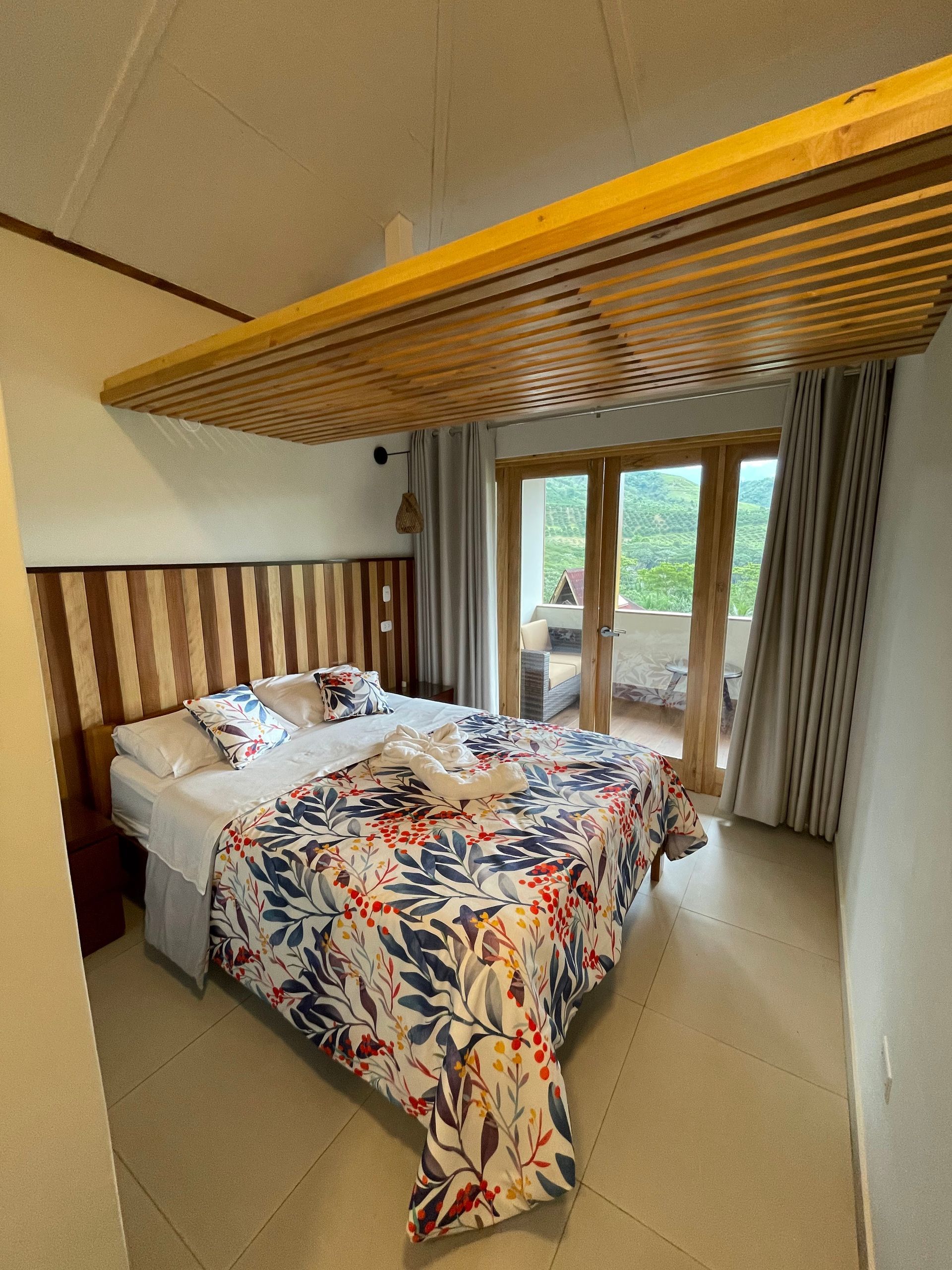 Dormitorio con cama, edredón floral, cabecera de madera, acceso al balcón y panel de techo de madera.
