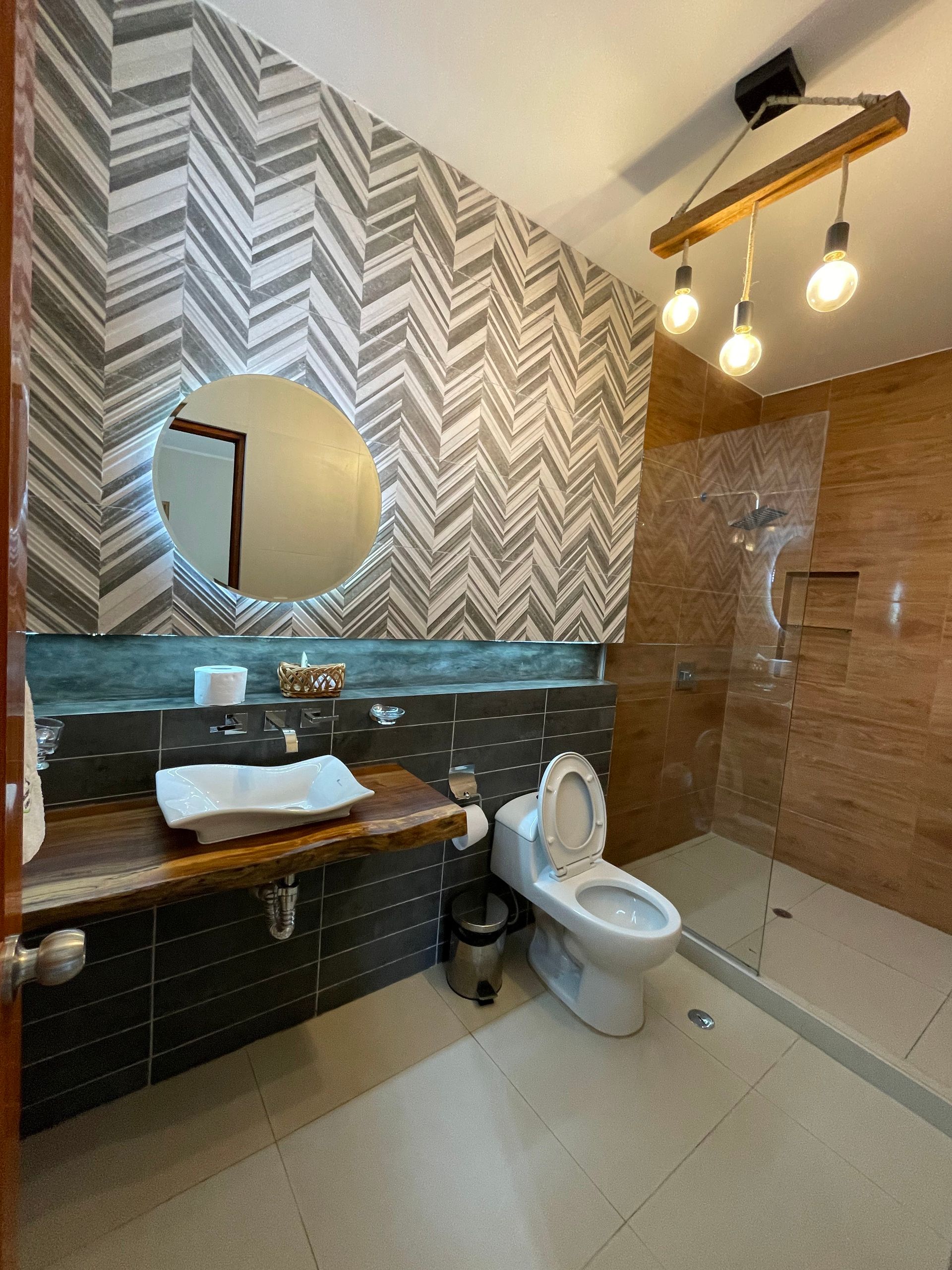 Baño con papel tapiz en zigzag, tocador de madera, espejo redondo, ducha de cristal e iluminación con bombillas expuestas.