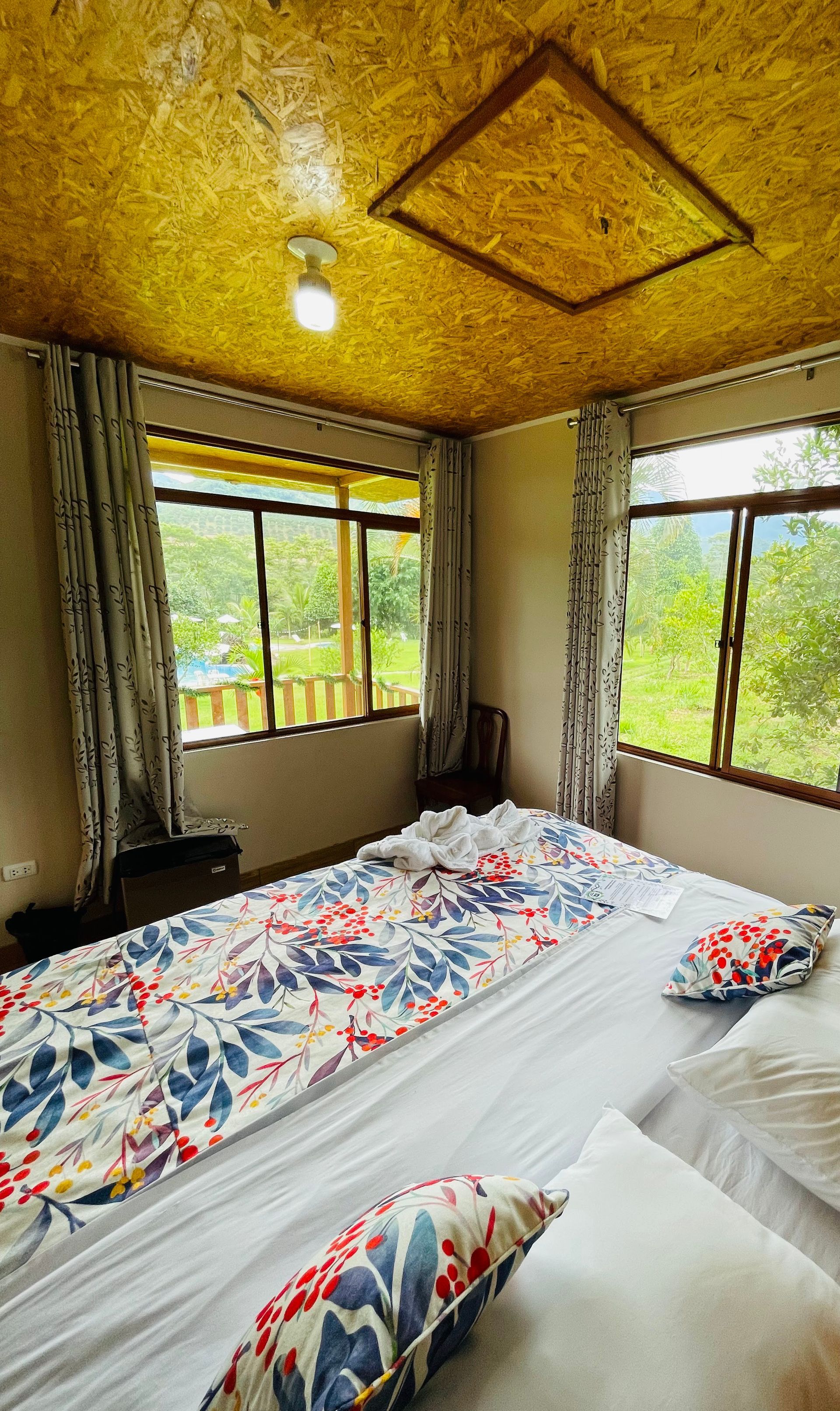 Un dormitorio con techo de OSB, sábanas con estampado floral y grandes ventanales con vistas a una zona verde y boscosa.