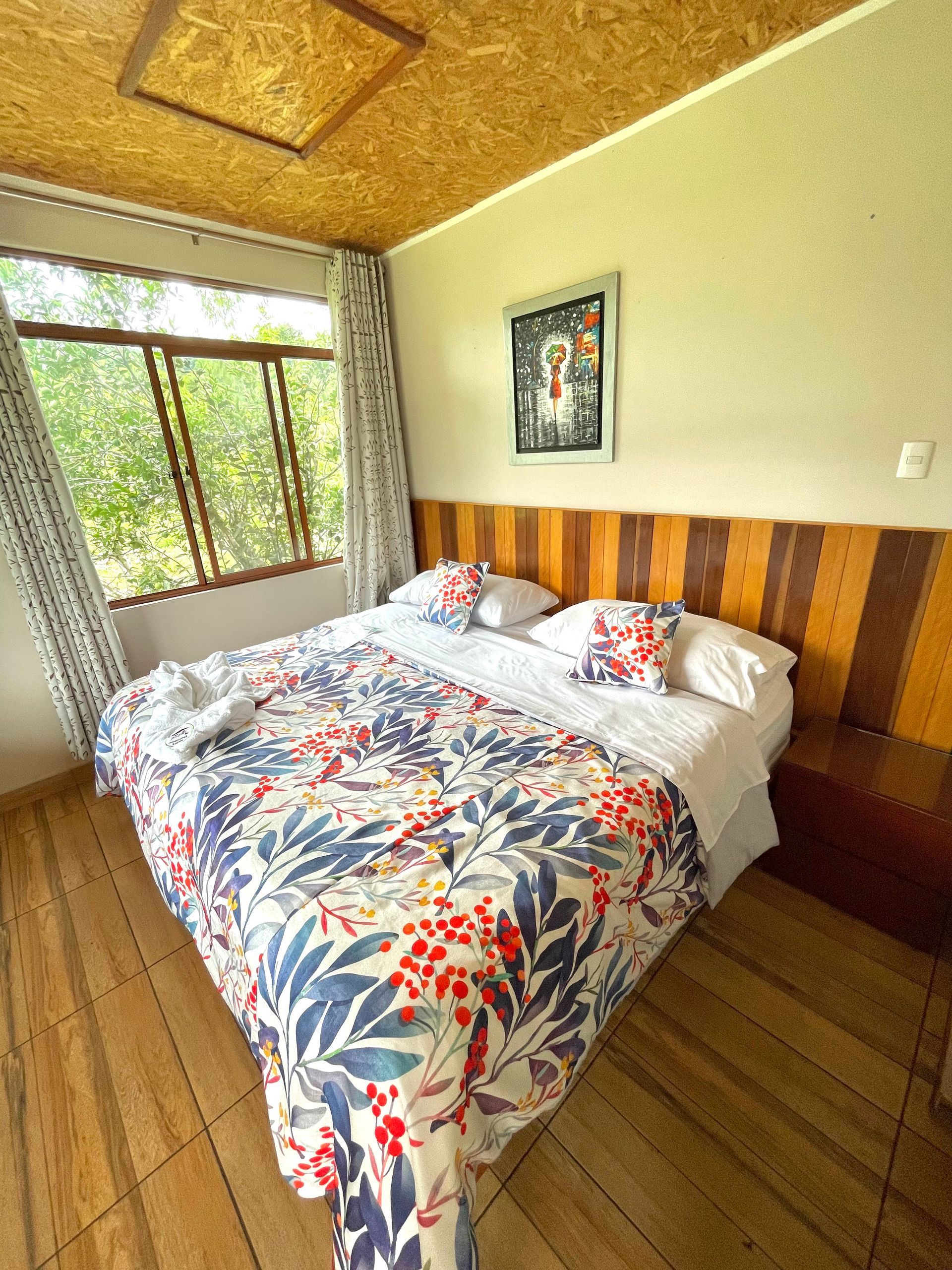 Un dormitorio acogedor con una colcha estampada en la cama, suelo y cabecero de madera, techo de OSB y una ventana.