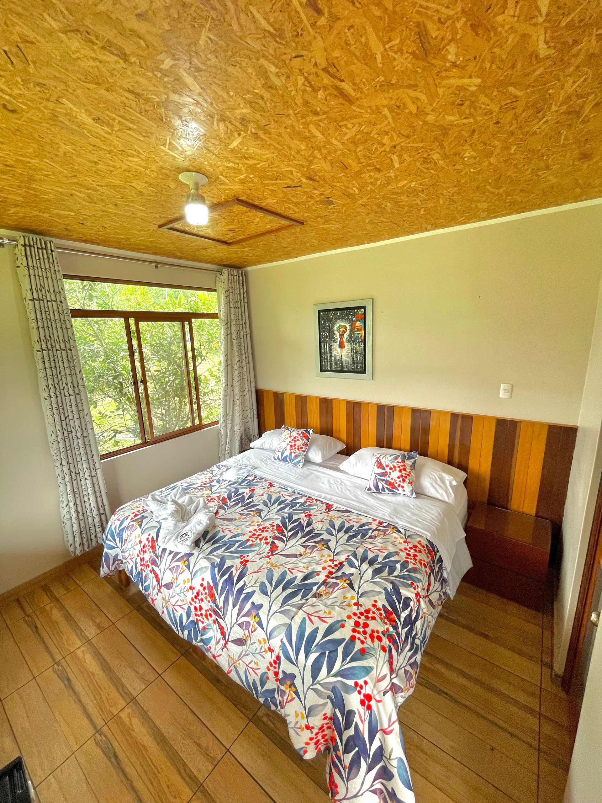 Un dormitorio con una colcha estampada, suelo de madera, techo de OSB y una gran ventana con vistas a la vegetación.