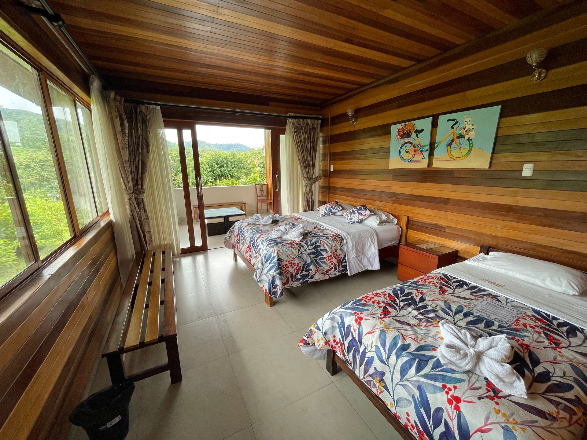 Un dormitorio rústico con paredes revestidas de madera, dos camas con ropa de cama estampada con flores y grandes ventanales.