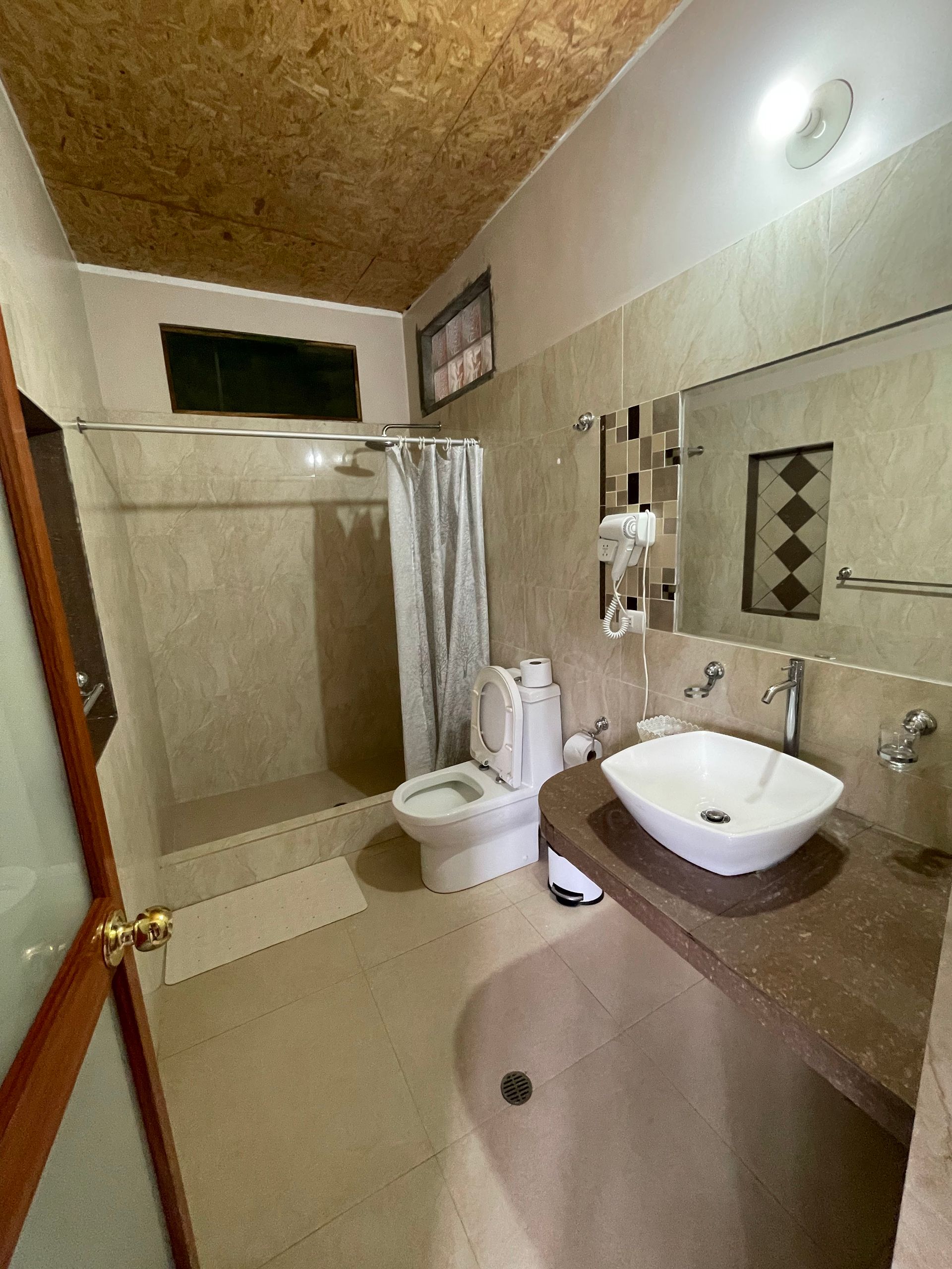 Un baño en tonos neutros que cuenta con una ducha a ras de suelo con cortina, un inodoro blanco y un lavabo.