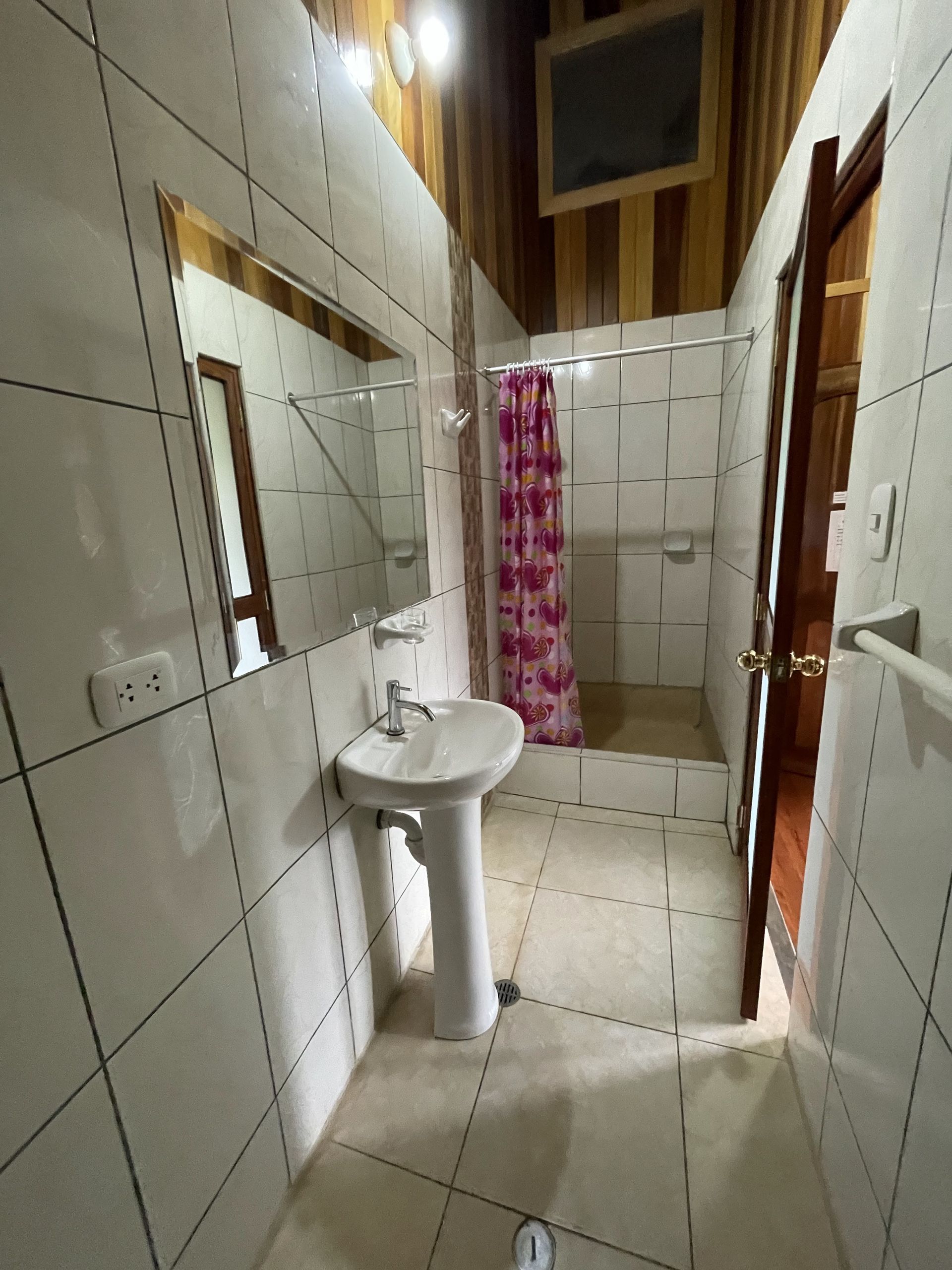 Un baño con paredes y suelos de azulejos blancos, un lavabo con pedestal y espejo, y una ducha a ras de suelo con cortina.