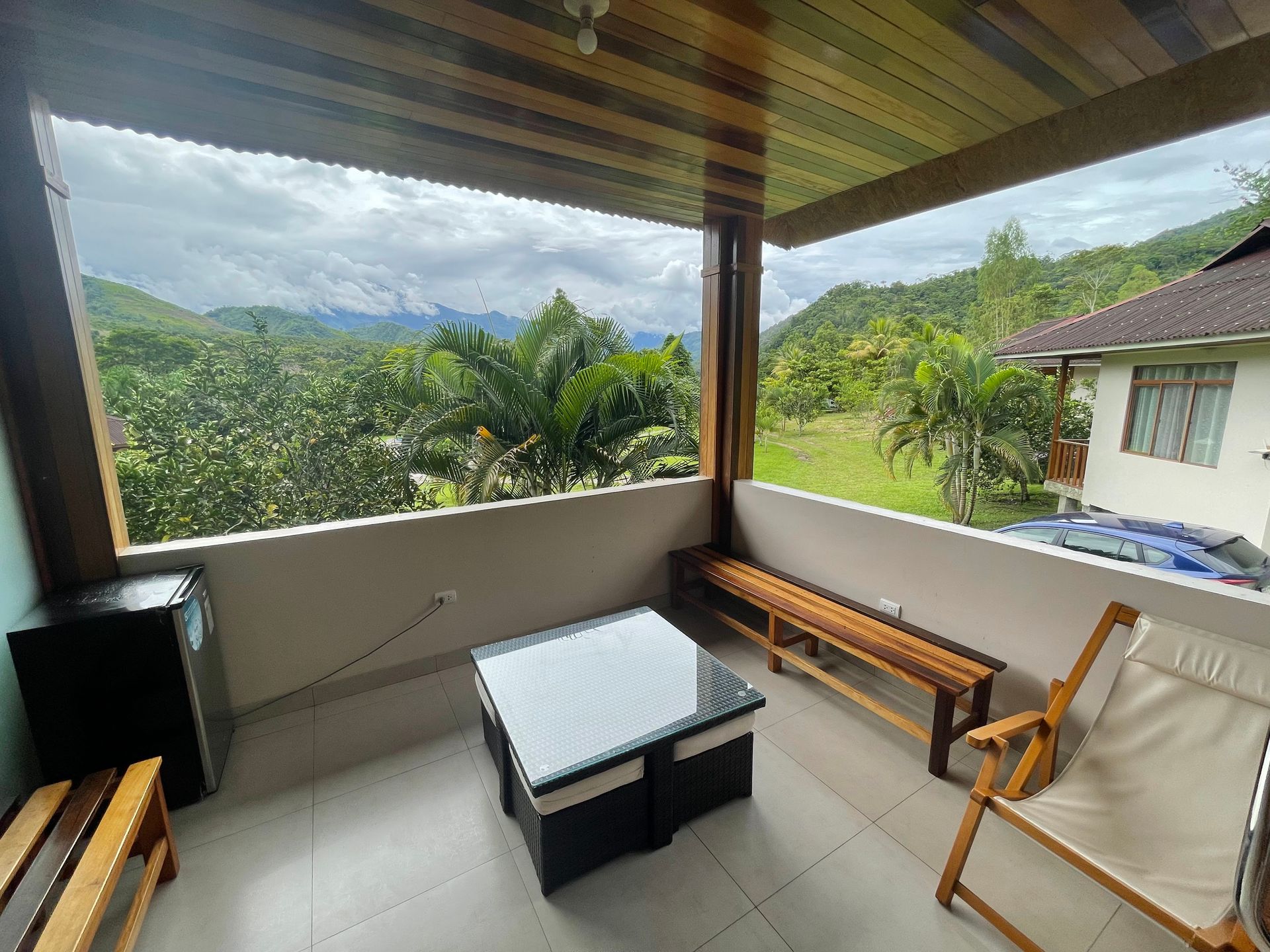 Un patio cubierto con mesa, bancos y sillas, con vistas a exuberantes montañas y vegetación bajo un cielo nublado.