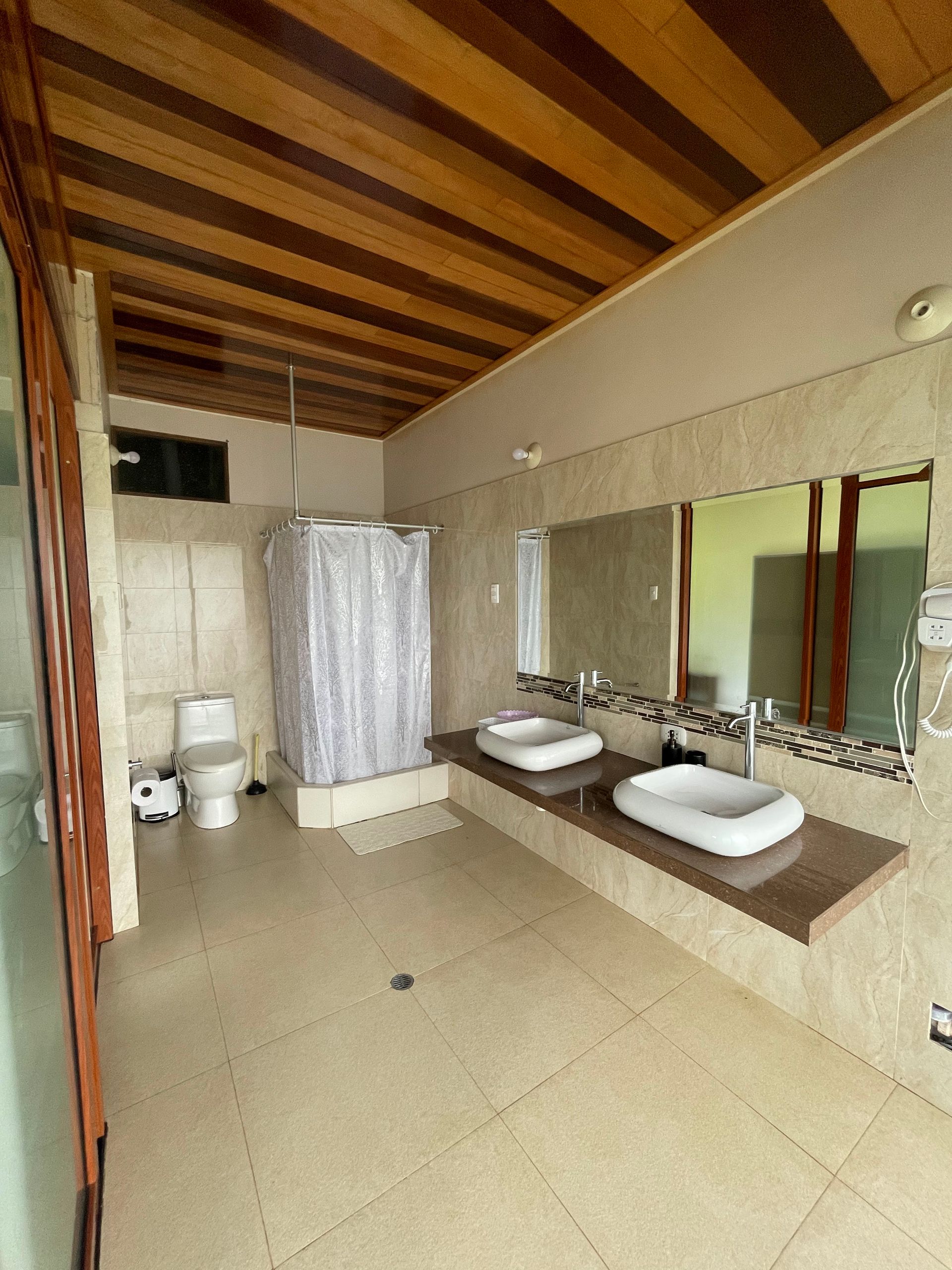 Un baño con tocador de doble lavabo, inodoro y ducha a ras de suelo con cortina, bajo un techo de madera inclinado.