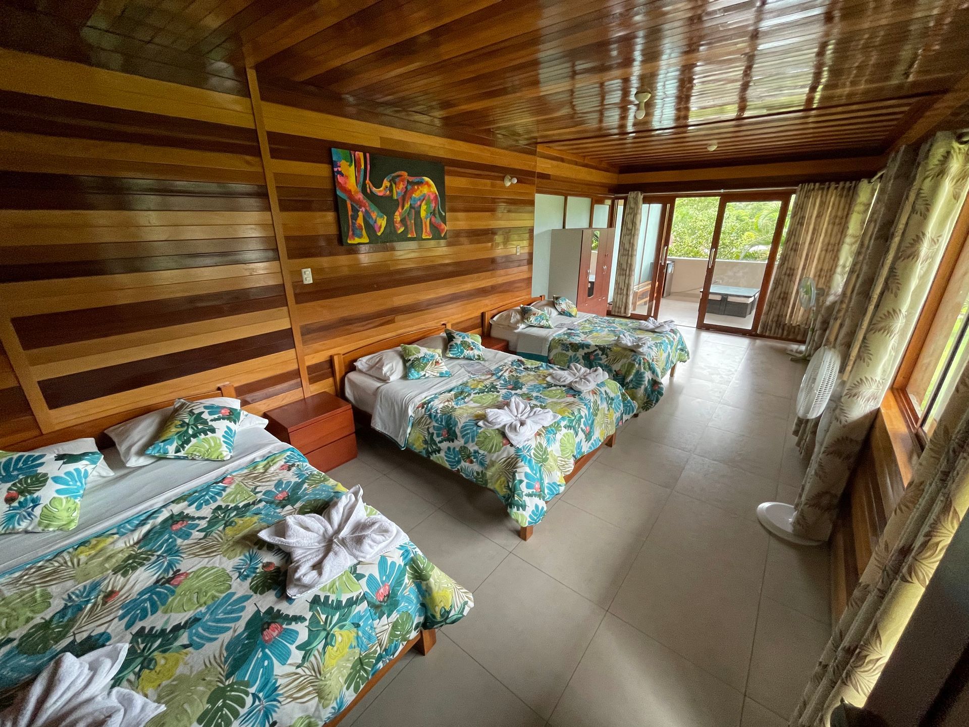 Un dormitorio con paredes revestidas de madera, tres camas con ropa de cama floral, suelos de baldosas y vistas al balcón.