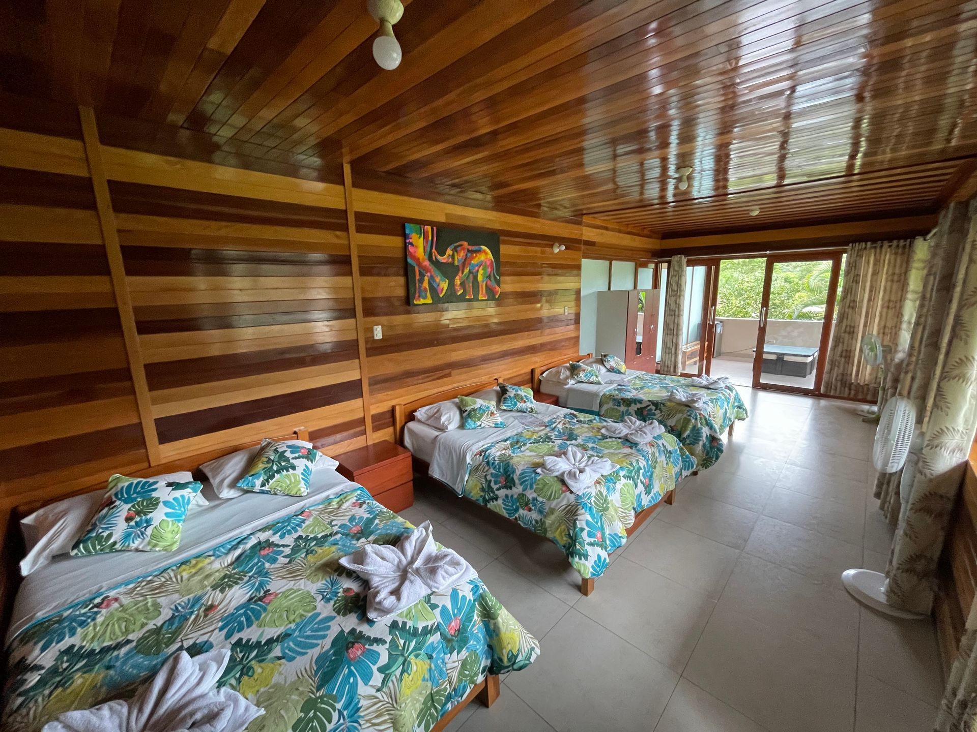Una habitación con tres camas, ropa de cama tropical en tonos verde azulado y blanco, paredes revestidas de madera.