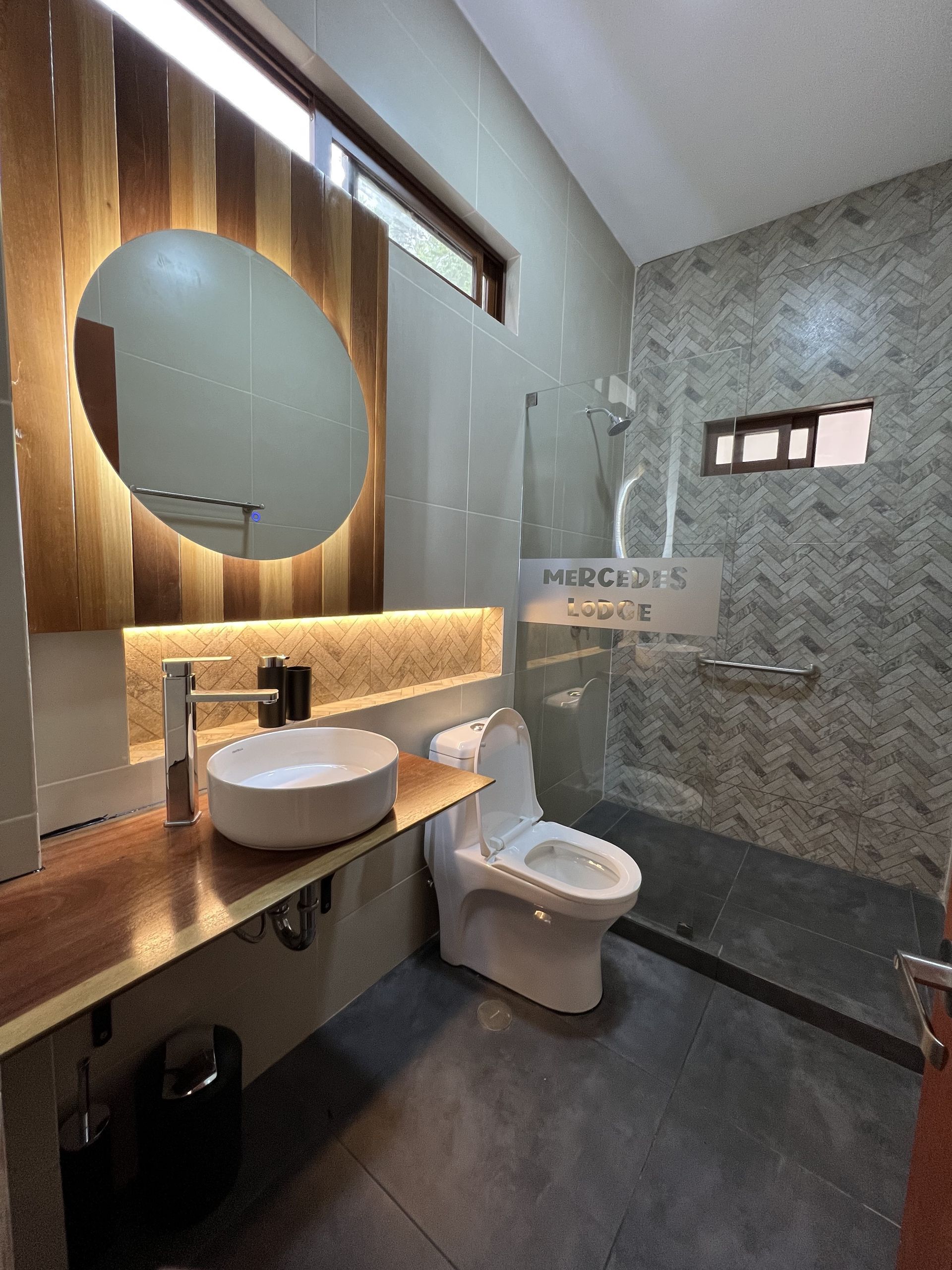 Un baño moderno con espejo circular con detalles de madera, lavabo sobre encimera y ducha con mampara de cristal 