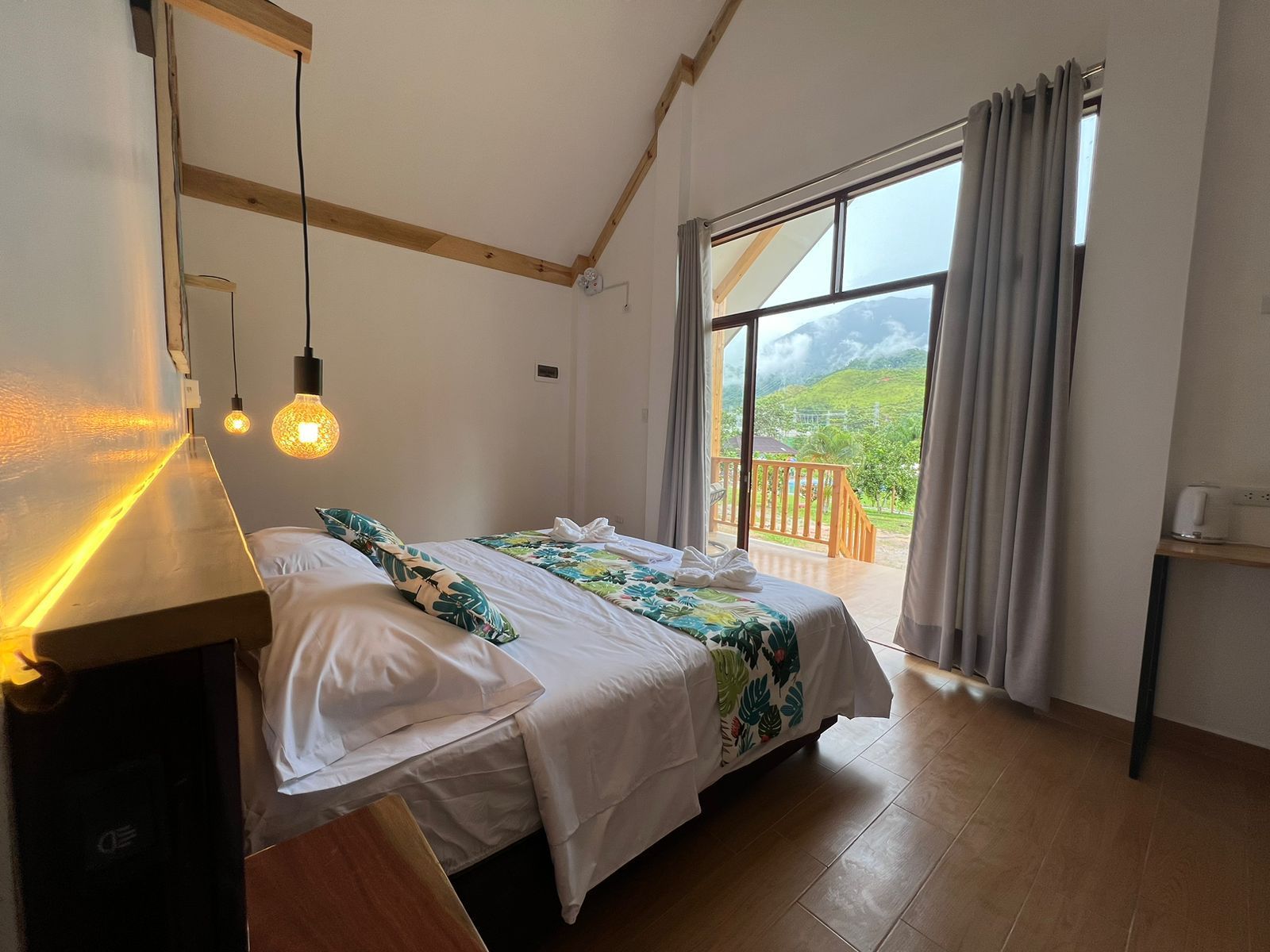 Una habitación de hotel moderna con cama de matrimonio, bombillas Edison colgantes, iluminación ambiental cálida y un balcón.