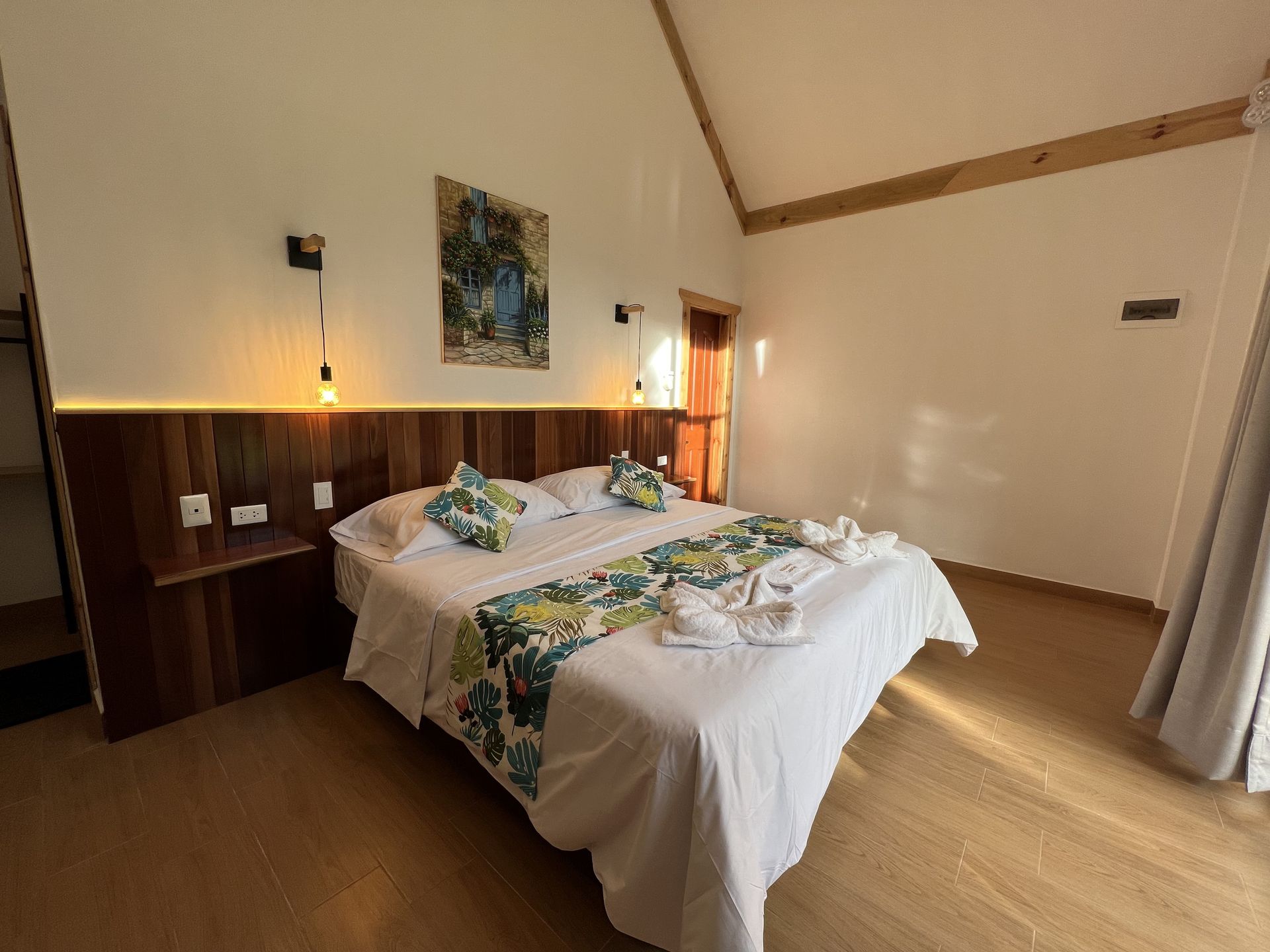 Un dormitorio con una cama que cuenta con un camino de mesa floral, sábanas blancas, cabecero de madera, iluminación.