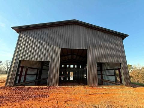 Gray metal barn with open doors on a red dirt lot under a blue sky.