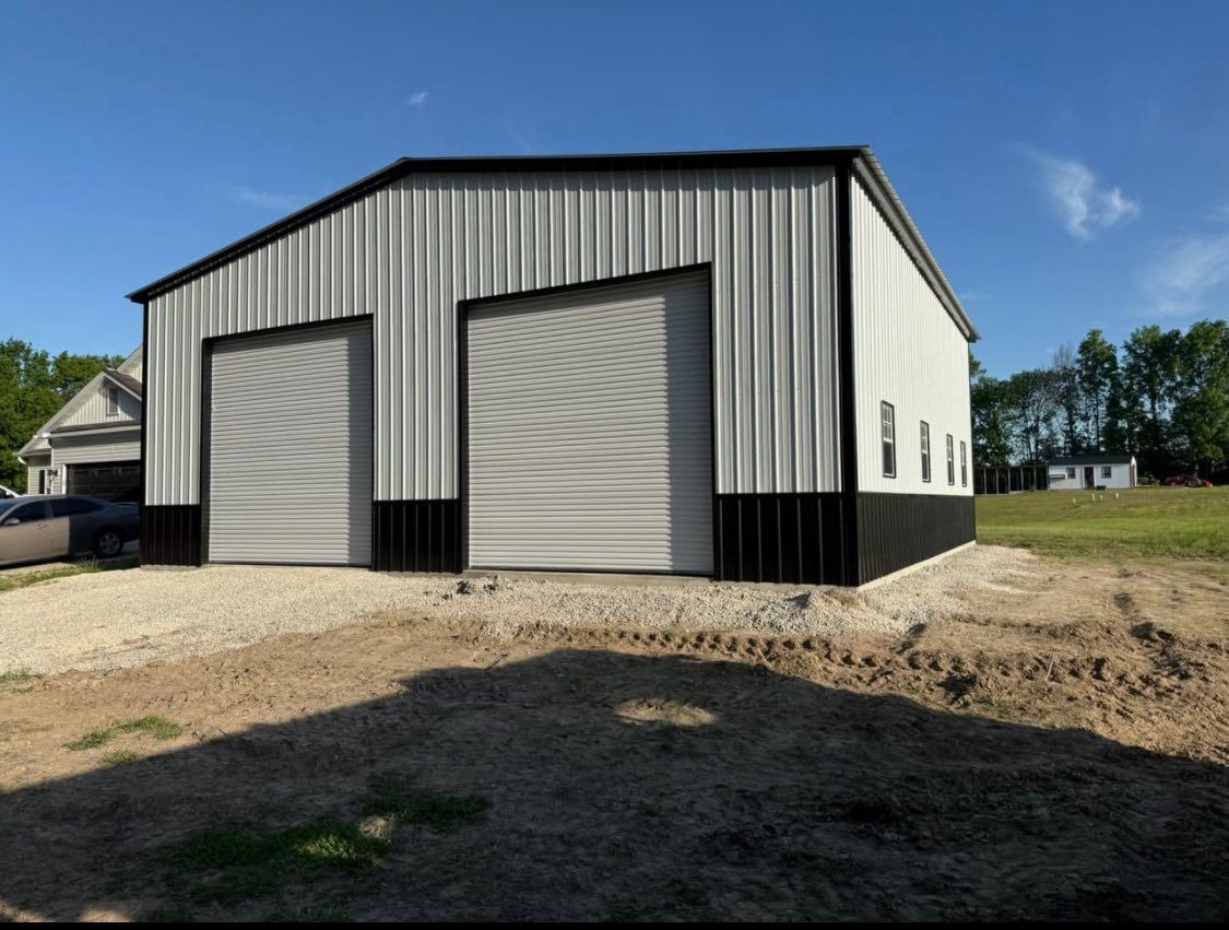 A metal workshop with two large garage doors, black accents, and a gravel foundation under a blue sky.