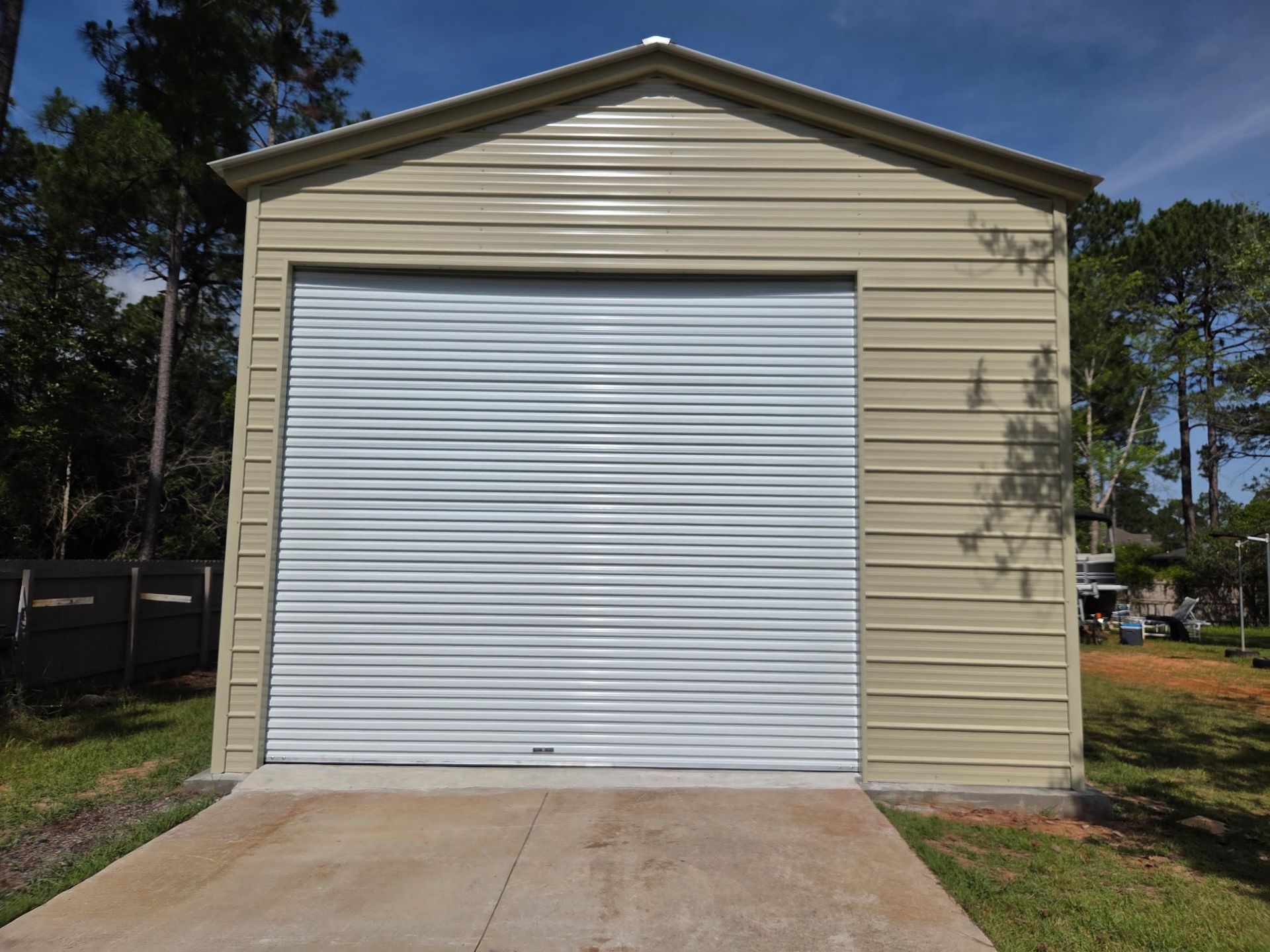 Tan metal garage with a white roll-up door. Concrete driveway in front, trees in the background.