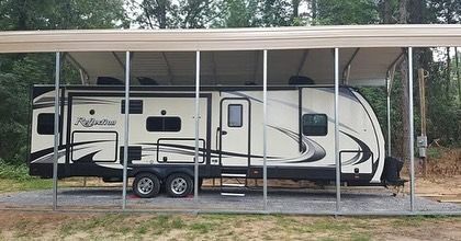 RV parked under a metal carport in a grassy area, surrounded by trees.