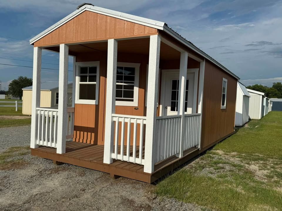 Brown and white cabin with porch, windows, and door, in a grassy field.