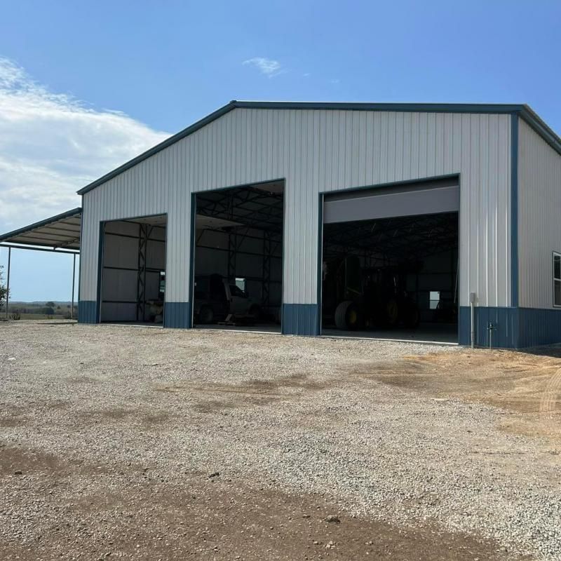 Large metal farm equipment shed with three open bays on a gravel lot under a clear blue sky
