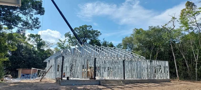 Metal frame house under construction, surrounded by trees, under a blue sky.