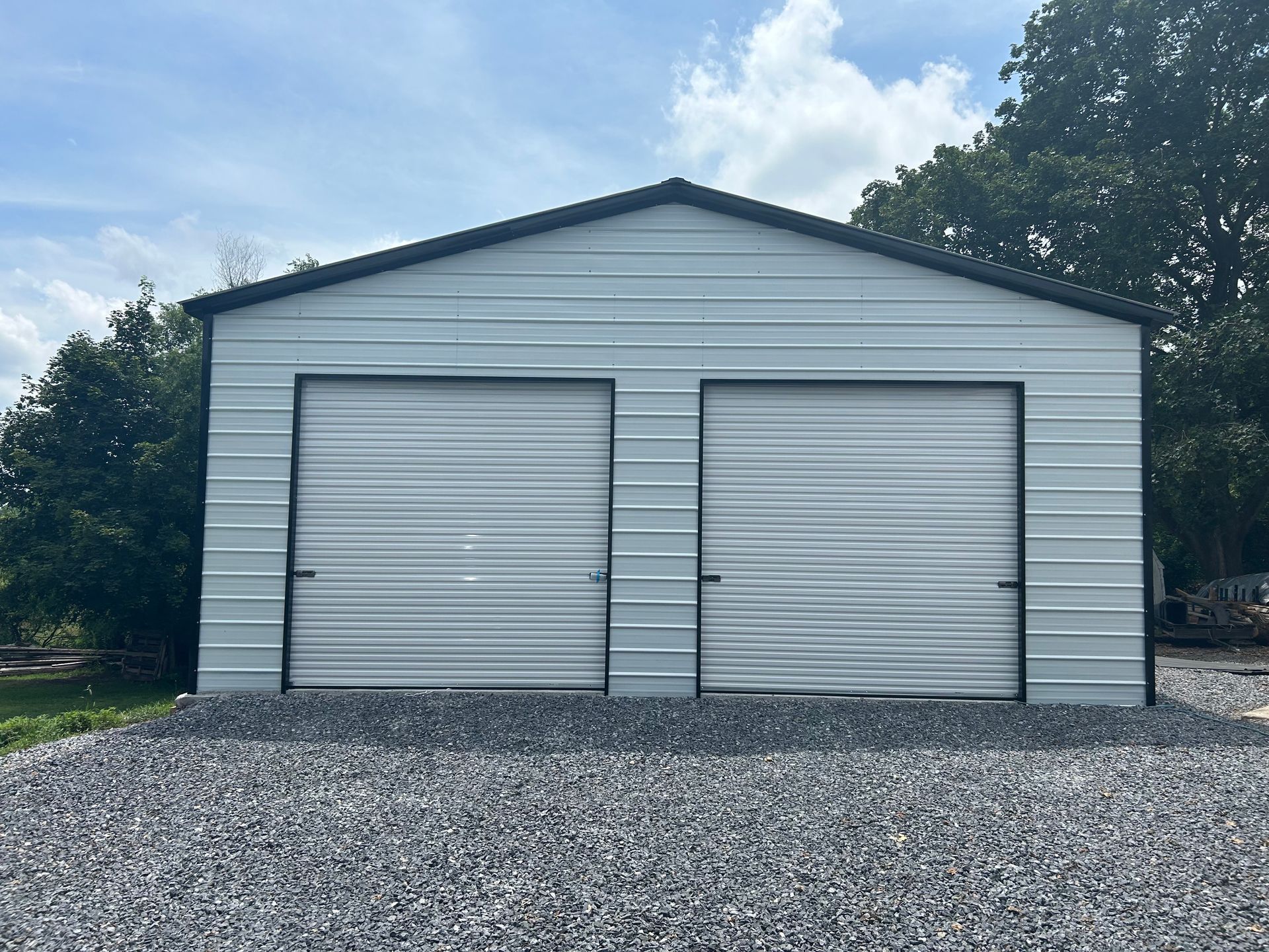 Gray metal two-car garage with black trim, gravel driveway, set against a backdrop of trees and sky.