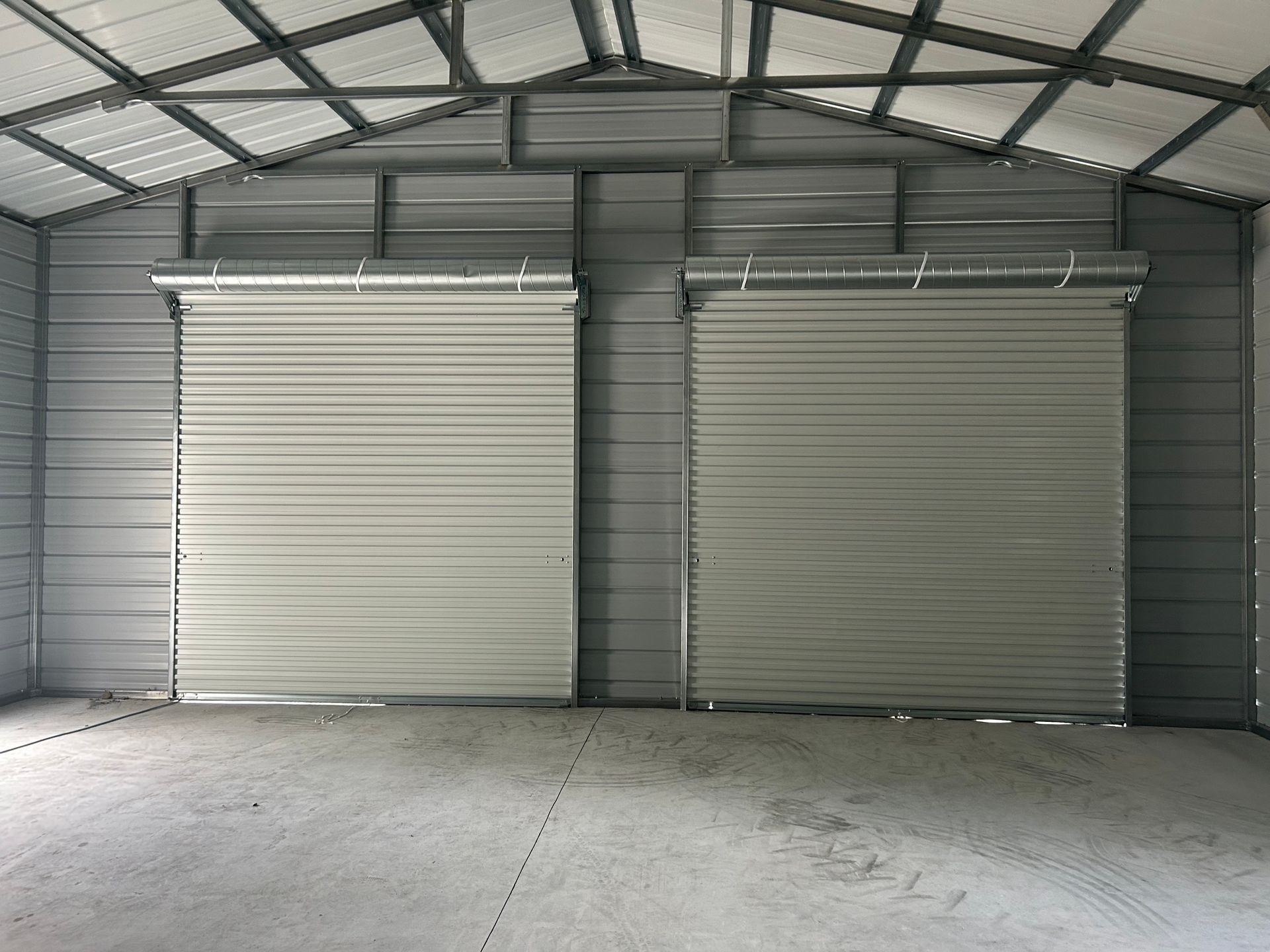 Inside a metal shed, two closed roll-up doors face the viewer; gray walls, roof, and concrete floor.