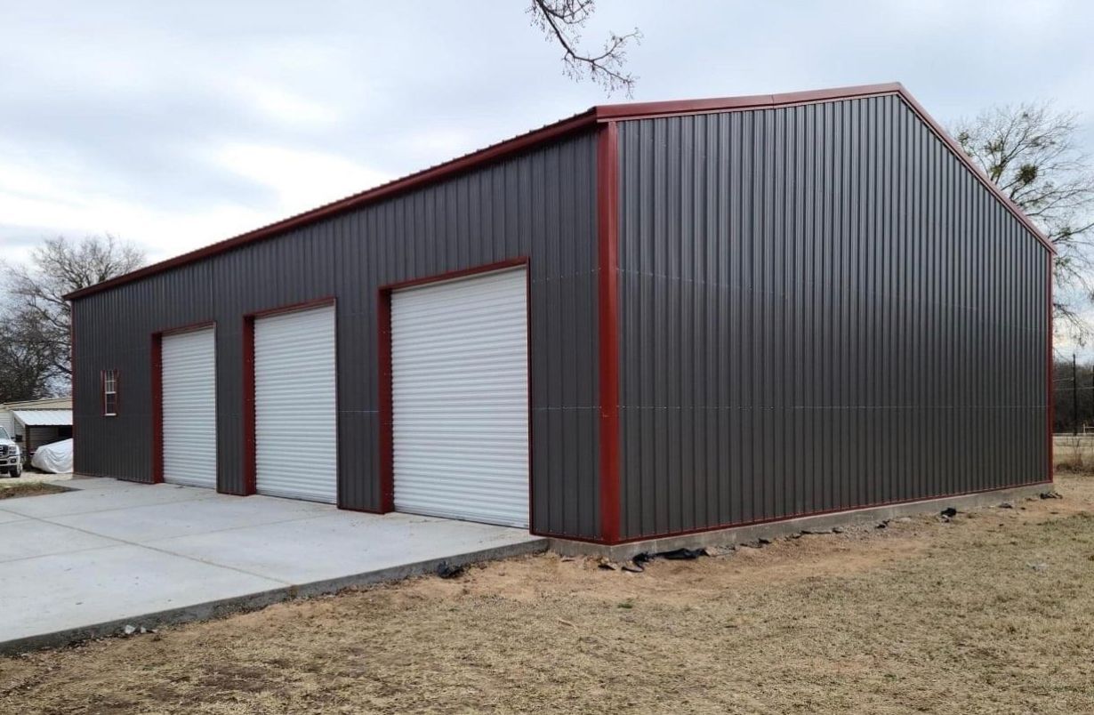 Gray metal building with three white garage doors, red trim, concrete slab, and brown grass.