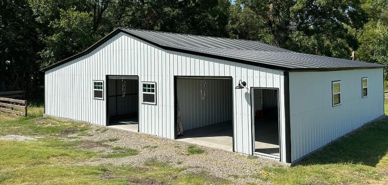 White metal building with black trim, two garage doors, and windows, set in a grassy area with trees in the background.