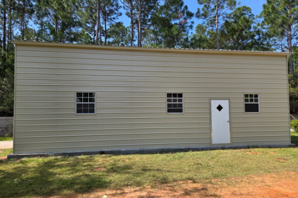 Beige metal building with small windows and a door, set in a grassy area with trees in the background.