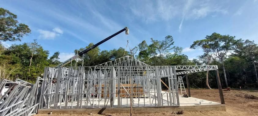 Construction site with steel frame structure being assembled by a crane, against a blue sky with trees.