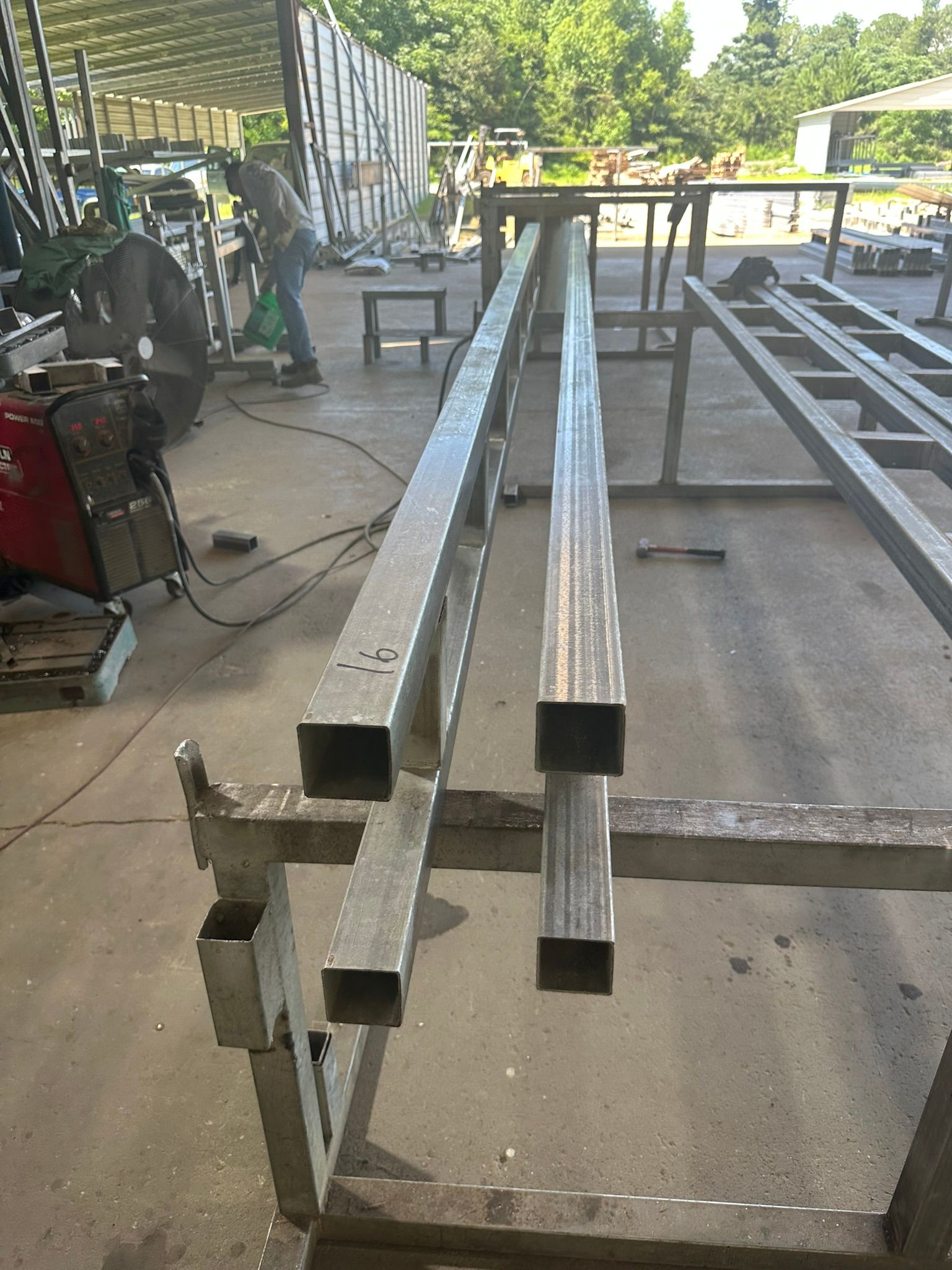 Two square metal tubes on a metal frame in a workshop; a worker is visible in the background.