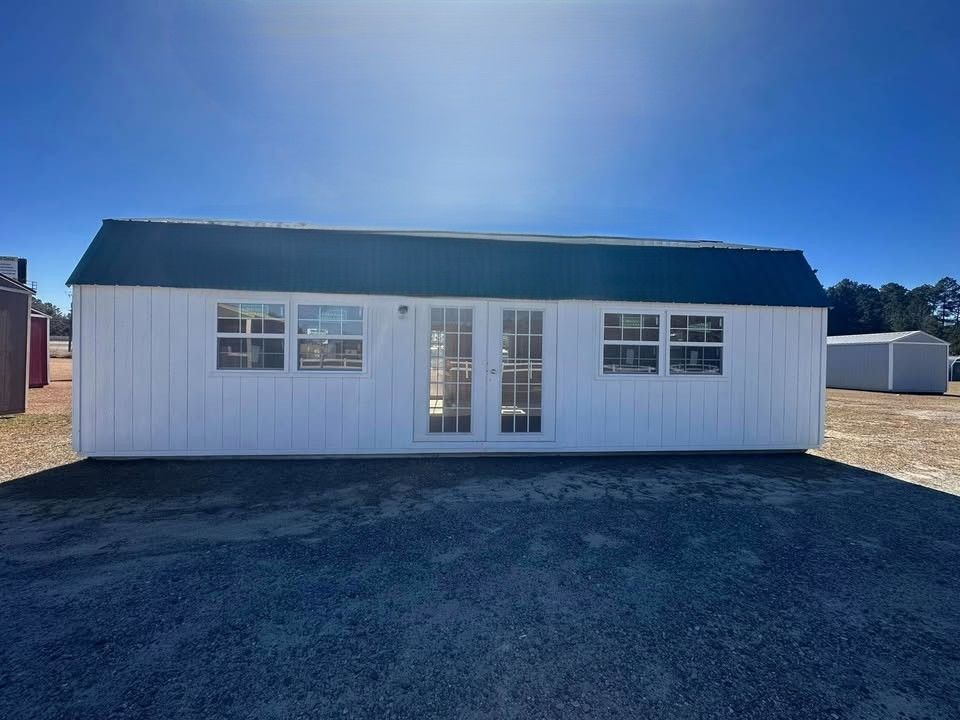 White shed with green roof, double doors, and windows on a sunny day.