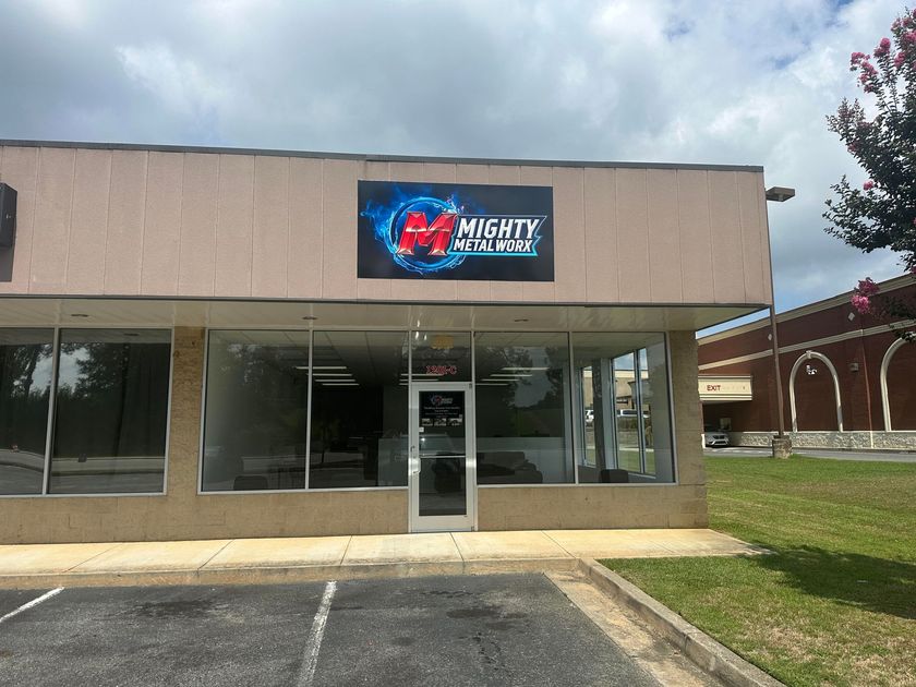 Mighty Comics storefront under a cloudy sky. Beige building with a sign. Open glass windows, door in the middle.