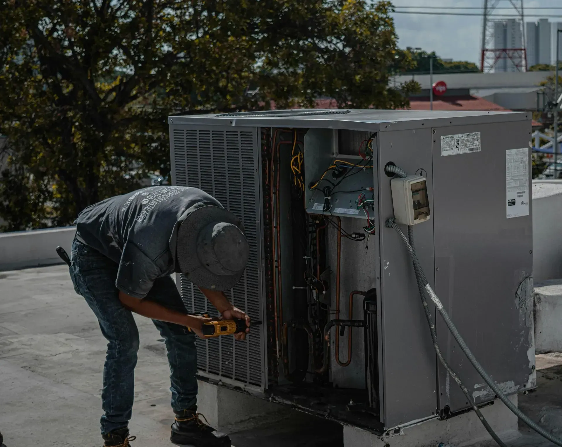 HVAC technician, wearing gray, repairing an air conditioning unit on a rooftop, using a screwdriver.