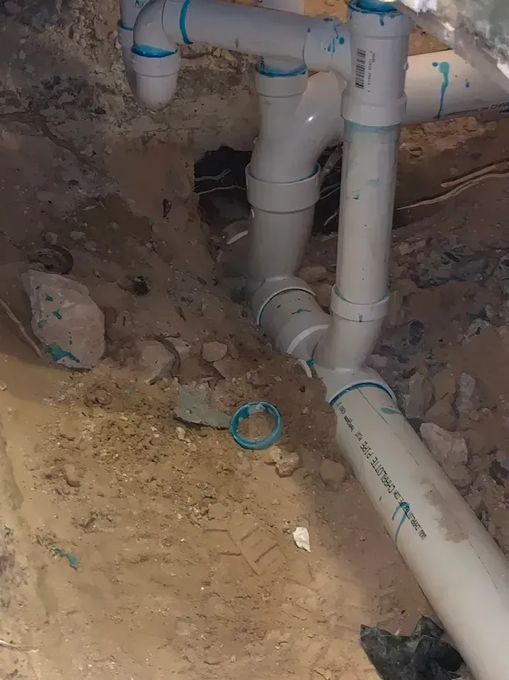 White PVC plumbing pipes in an excavated dirt area. Pipes connect at angles, with a hole in the background.