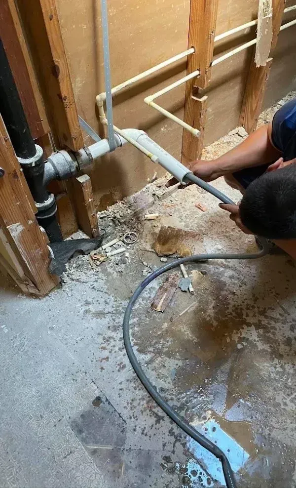 Plumber working on exposed pipes in a bathroom with tools and water present.