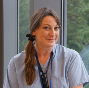 A woman in a blue scrub top with a lanyard around her neck smiles for the camera